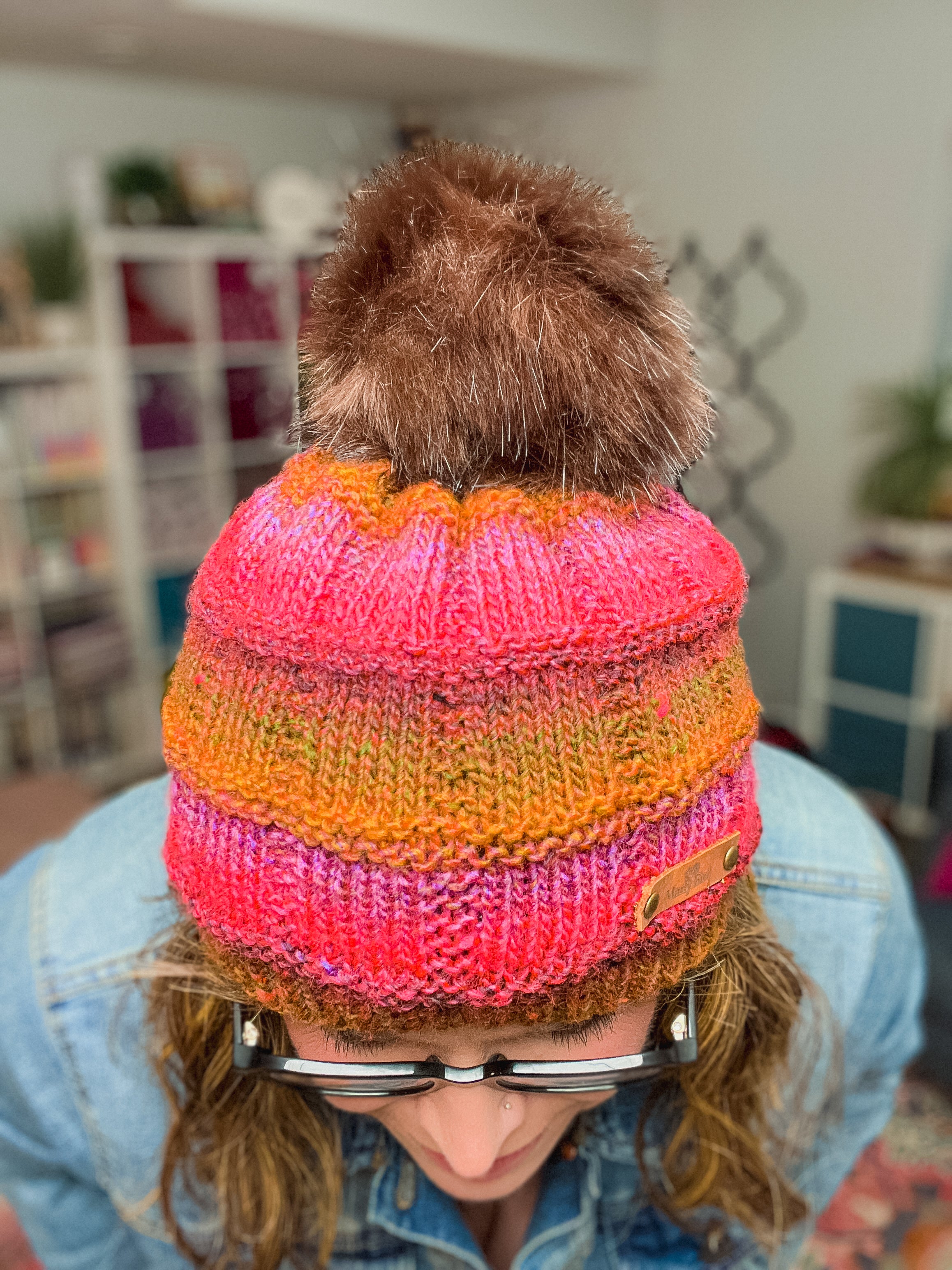 Someone wearing a Rory Knit Hat made from the Textured Knit Beanie PDF pattern (Toddler to Adult), in bright pink, orange, and yellow with a brown faux fur pom-pom, pairs it with glasses and a denim jacket indoors while looking down.