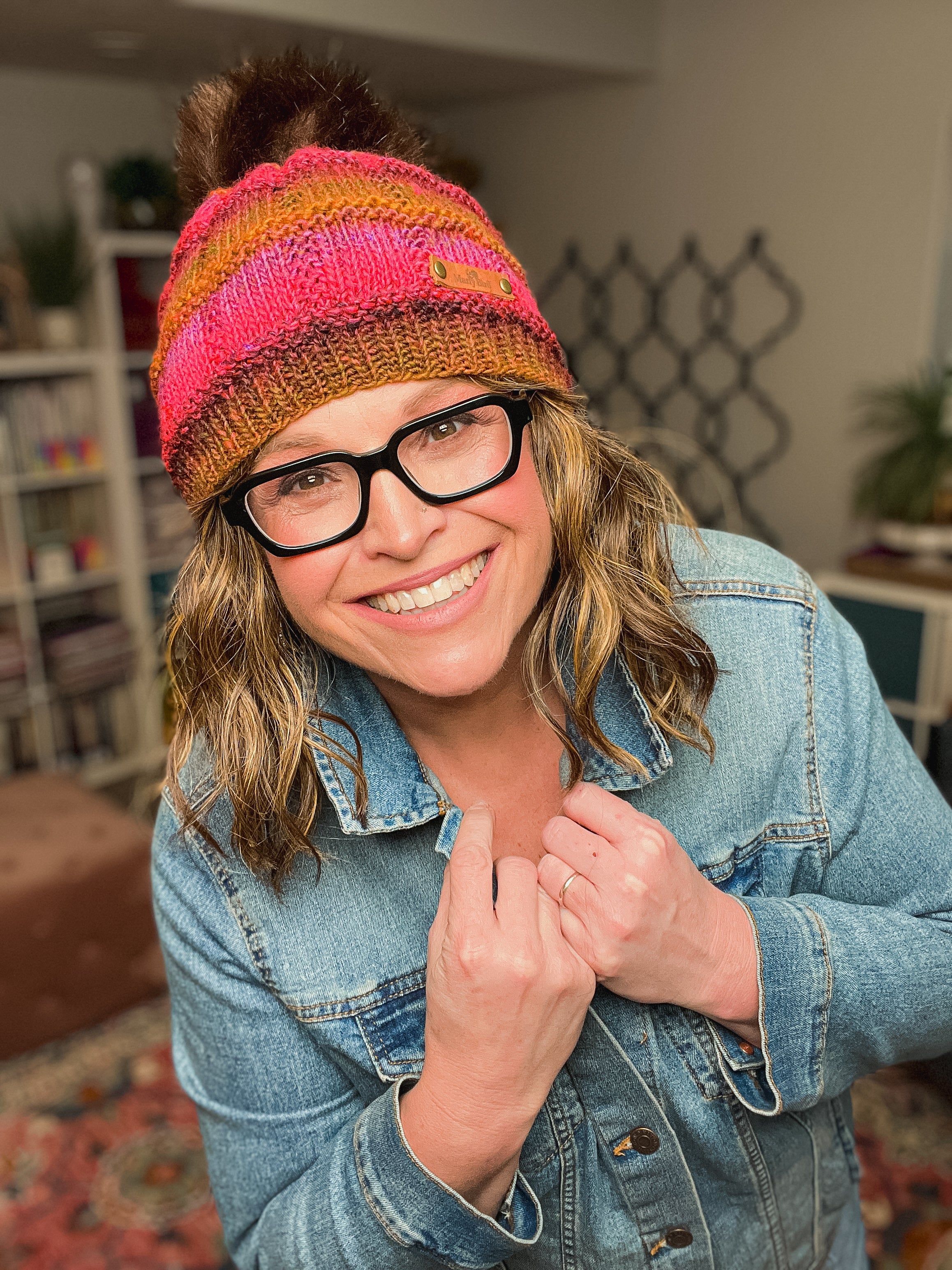 A smiling person in glasses wears a colorful Rory Knit Hat Pattern – Textured Knit Beanie (PDF, Toddler to Adult) with a pom-pom and a denim jacket indoors, holding the jacket collar. Books and plants are visible on shelves in the background.