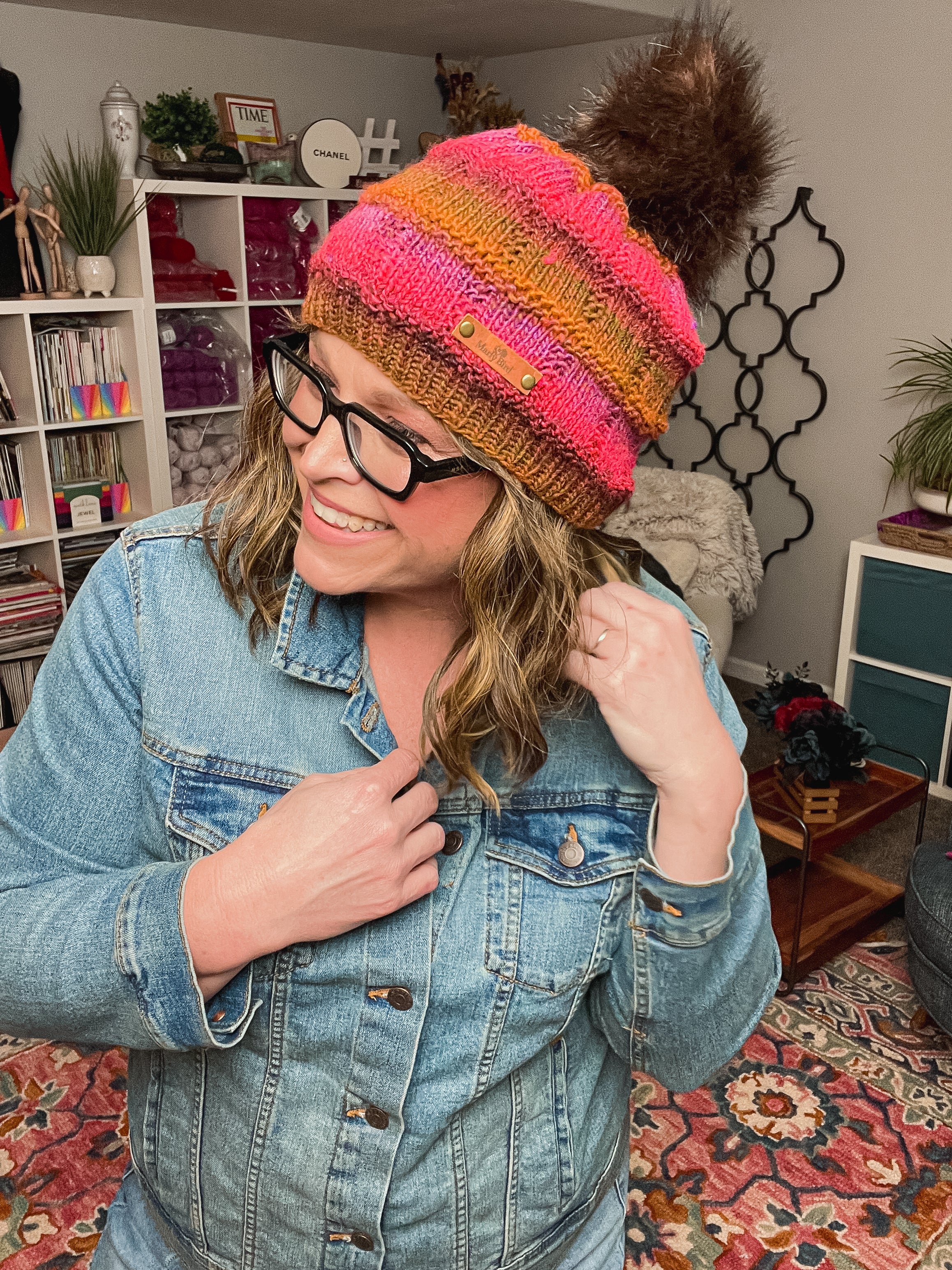 A woman in a denim jacket and glasses smiles while wearing the Rory Knit Hat, a colorful textured beanie with a brown pom-pom. She stands in a cozy room with shelves, plants, and patterned rugs. PDF pattern fits toddler to adult.