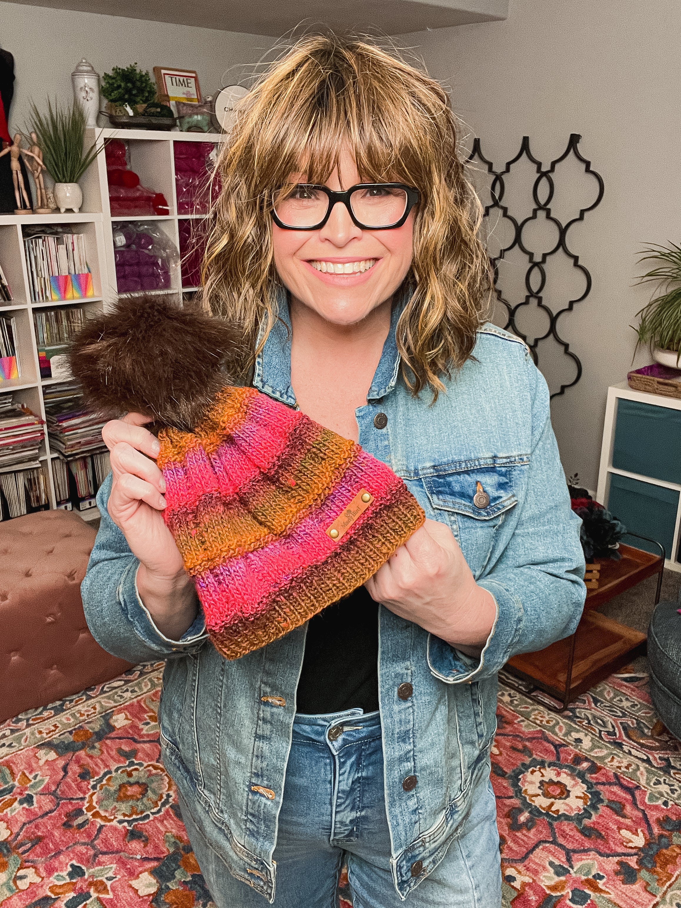 A smiling woman with wavy hair and glasses holds up a colorful Rory Knit Hat, made using the Textured Knit Beanie PDF pattern (toddler to adult). She wears a denim jacket in a cozy, vibrant room with a patterned rug.
