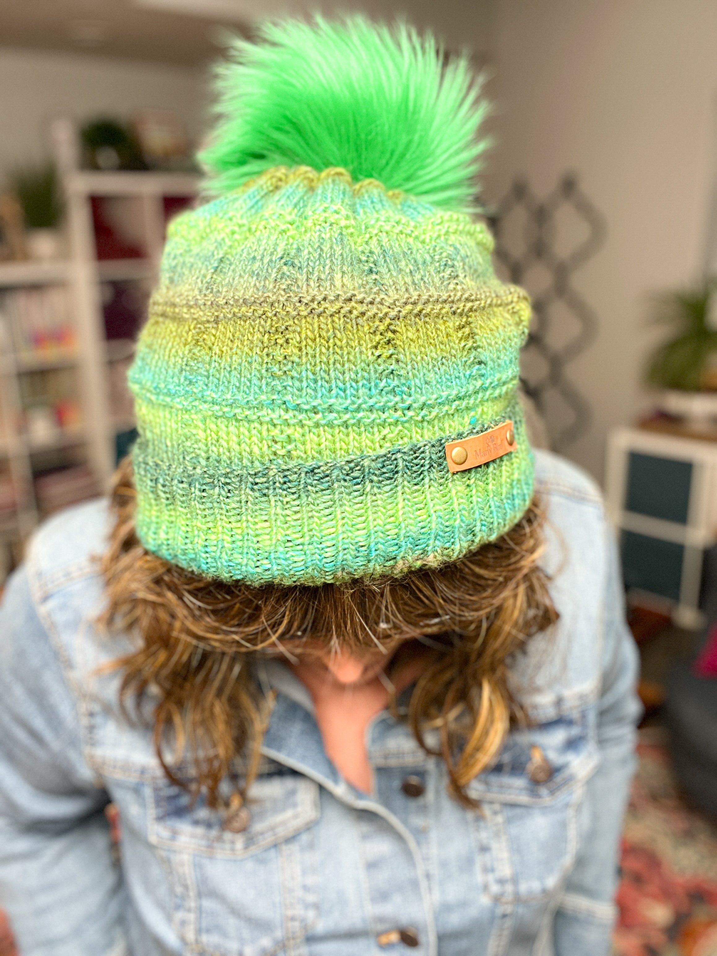 Someone wearing a green and blue Rory Knit Hat, made from the Textured Knit Beanie PDF pattern (Toddler to Adult), looks down as wavy brown hair escapes the cozy beanie. They're in a light denim jacket, with bookshelves and houseplants behind them.