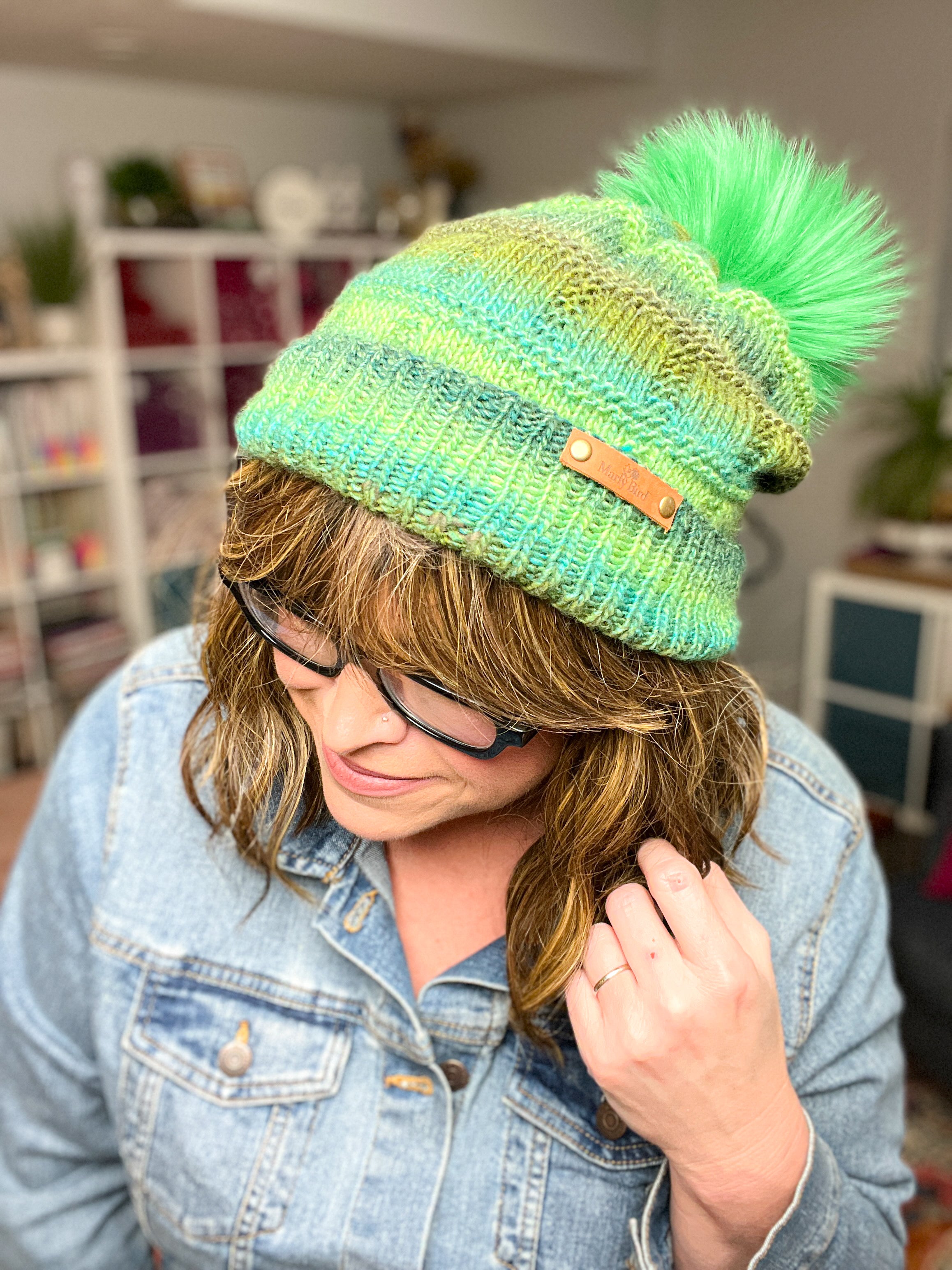 A woman in glasses and a denim jacket smiles, modeling the bright green Rory Knit Hat—a textured, worsted weight knit beanie with a large matching pom-pom. Bookshelves and plants create a cozy backdrop. Pattern available as PDF (Toddler to Adult).