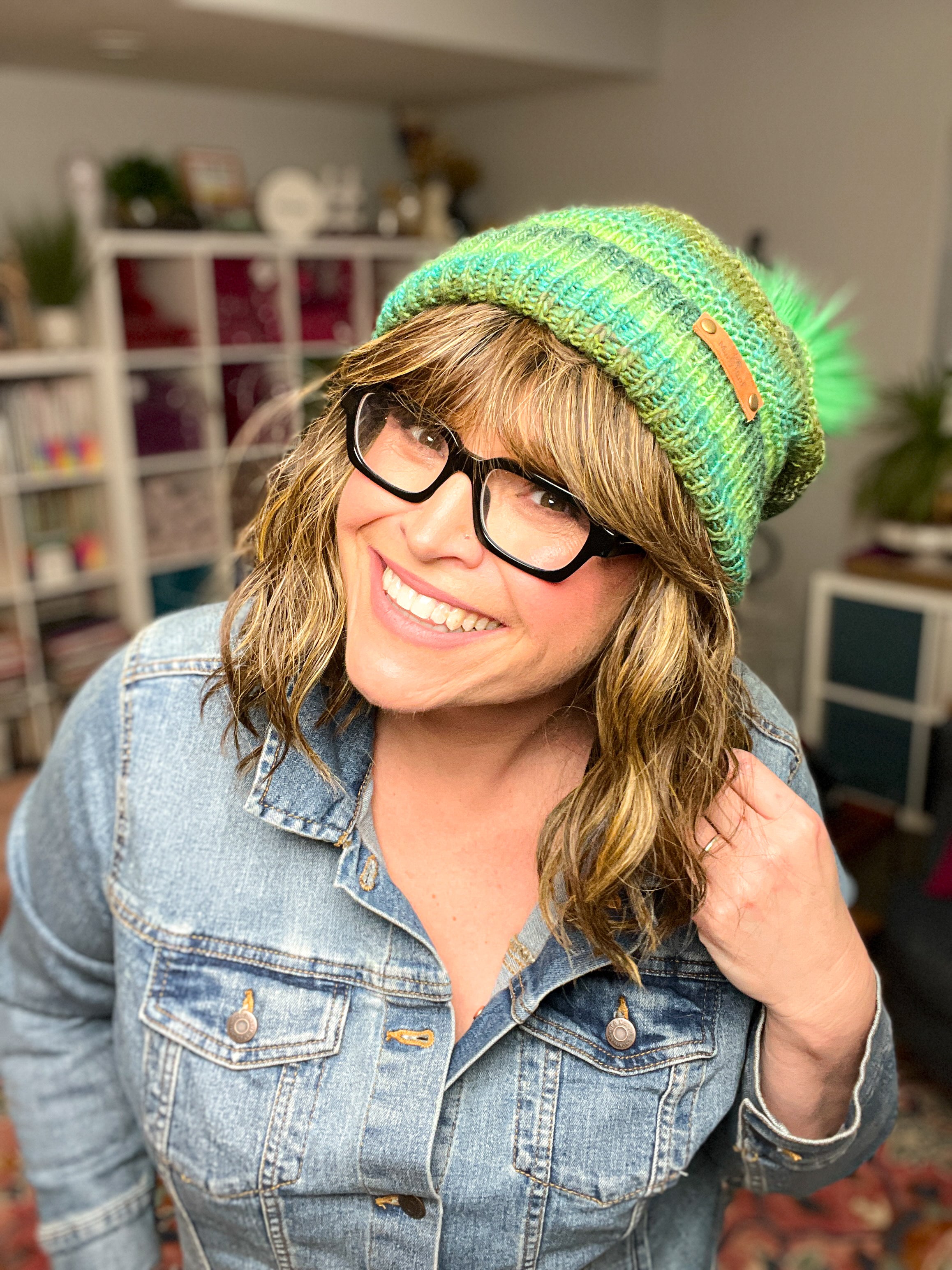 A smiling woman with wavy hair and glasses wears a denim jacket and the Rory Knit Hat—a green textured beanie made from the knit PDF pattern—while standing indoors with bookshelves and decor in the background.