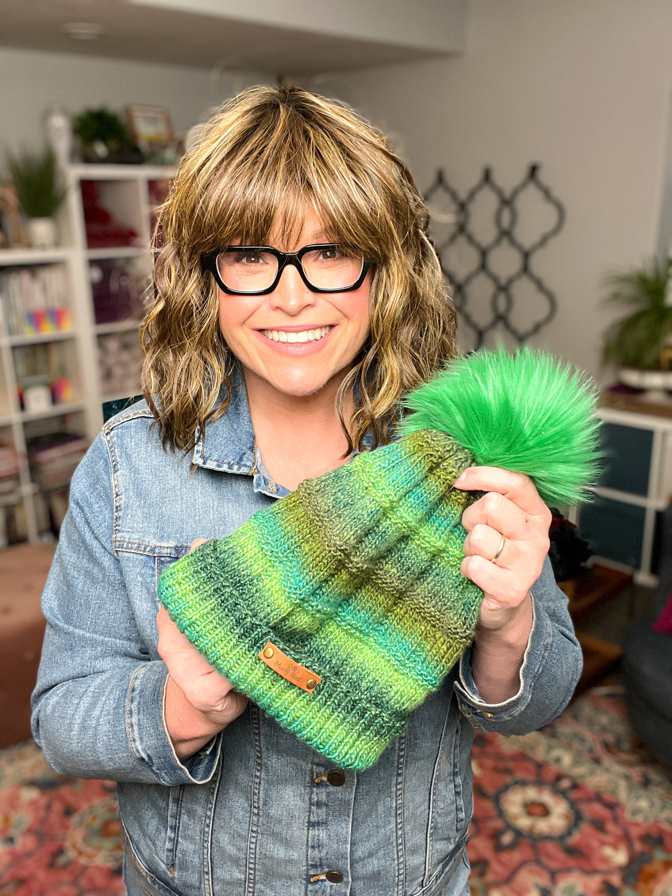 A smiling person with wavy hair and glasses holds the Rory Knit Hat—a green and blue textured knit beanie with a large green pom-pom. Wearing a denim jacket, they proudly showcase the Rory Knit Hat Pattern PDF (Toddler to Adult) in a cozy, well-lit room.