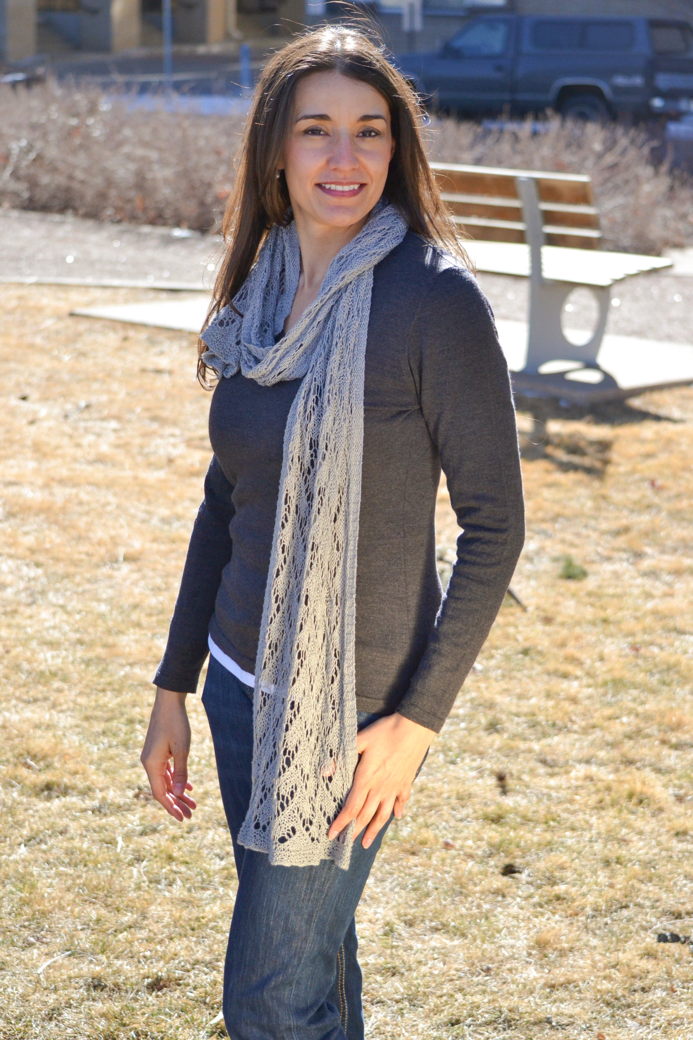 A woman smiles outdoors on dry grass, wearing a dark long-sleeve shirt, blue jeans, and the Shangri-La Knit Shawl Pattern—a lightweight lace shawl. A bench and bushes are visible in the background.