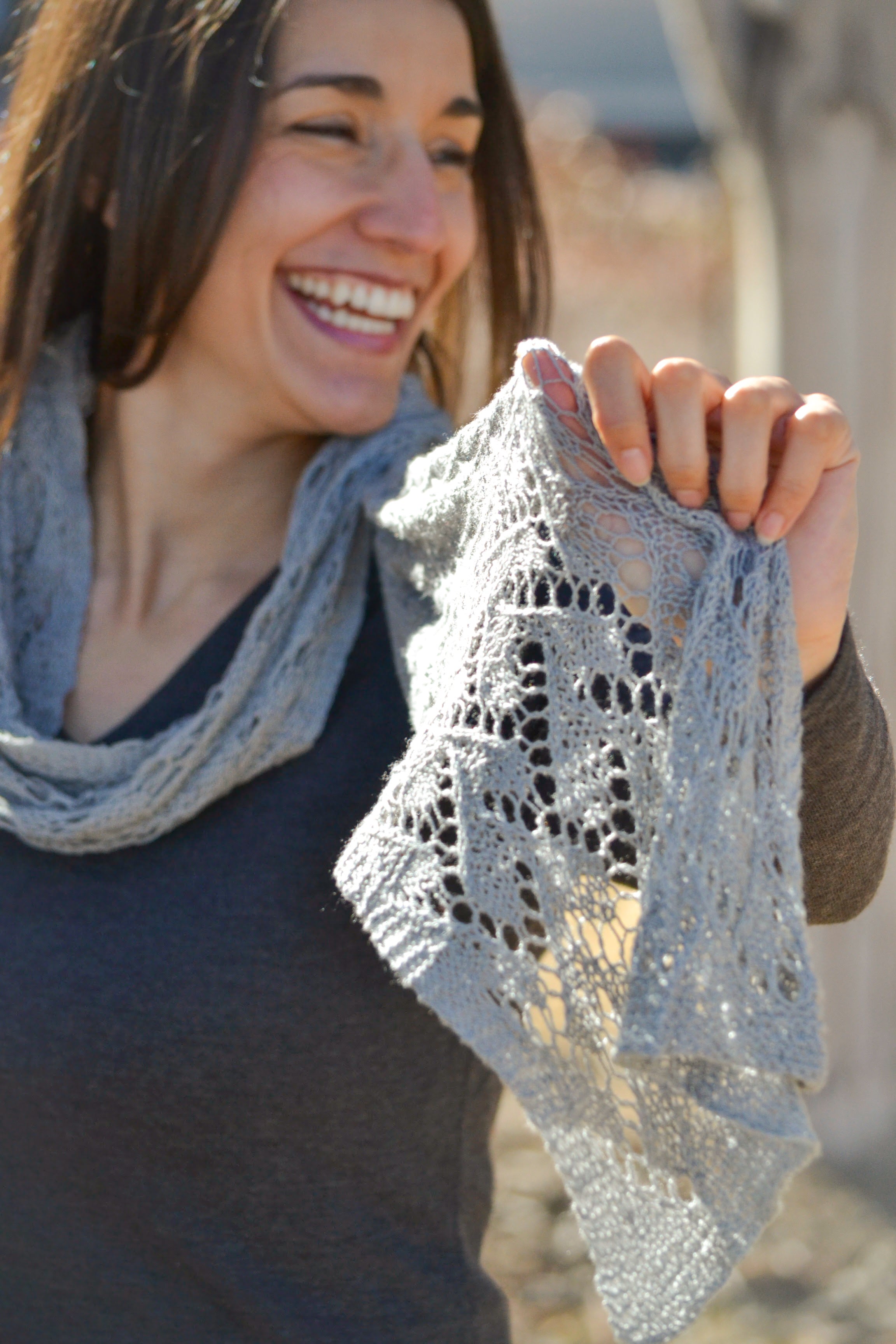 A smiling woman with long brown hair, wearing a dark top, holds up the Shangri-La Knit Shawl Pattern—a light gray, lightweight lace shawl (scarf or wrap) knitting PDF—showing its intricate design outdoors in bright daylight.