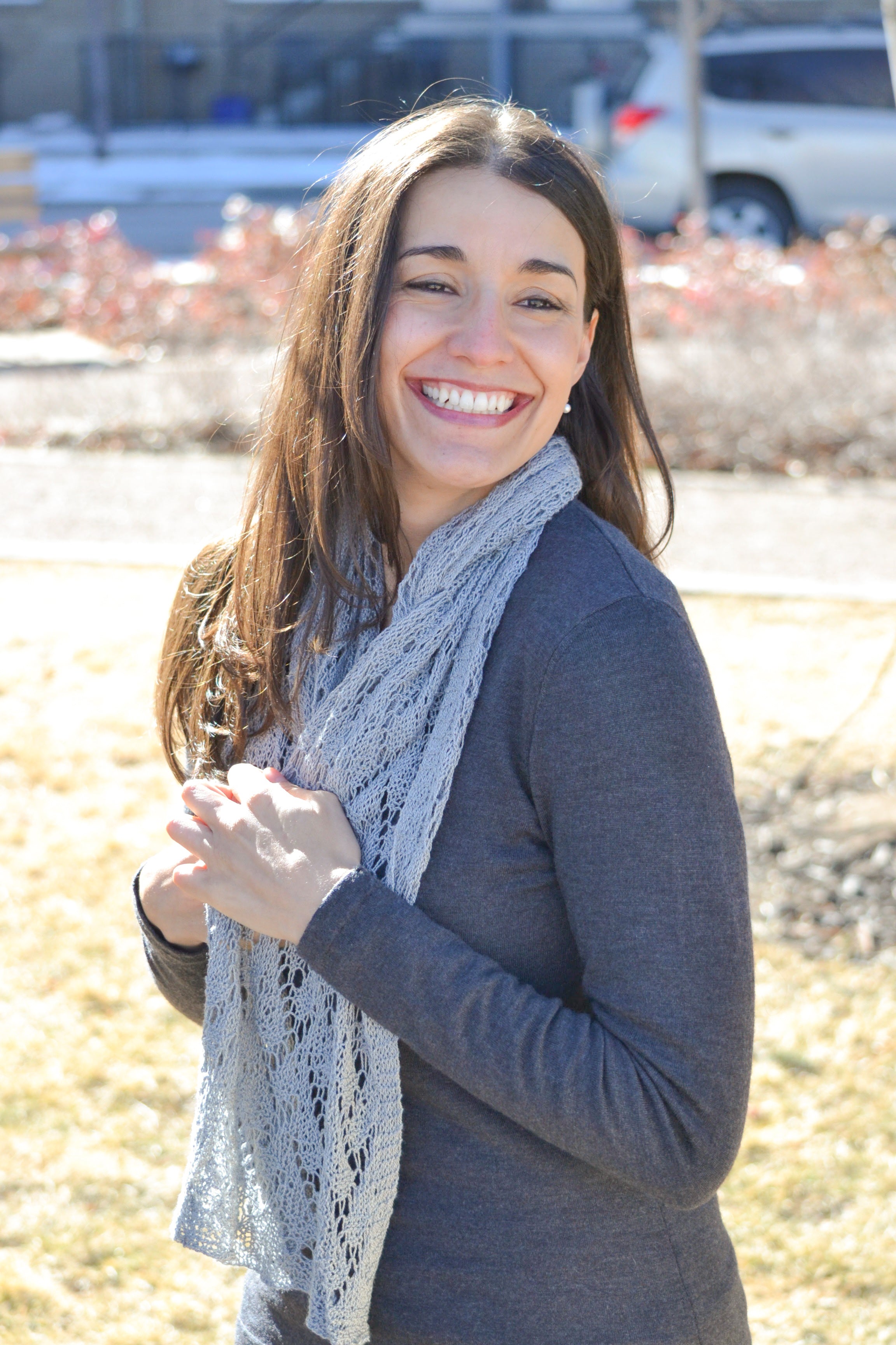 A woman with long brown hair stands outdoors on a sunny day, smiling brightly while wearing a dark sweater and the Shangri-La Knit Shawl made from the lightweight lace shawl knitting PDF pattern.