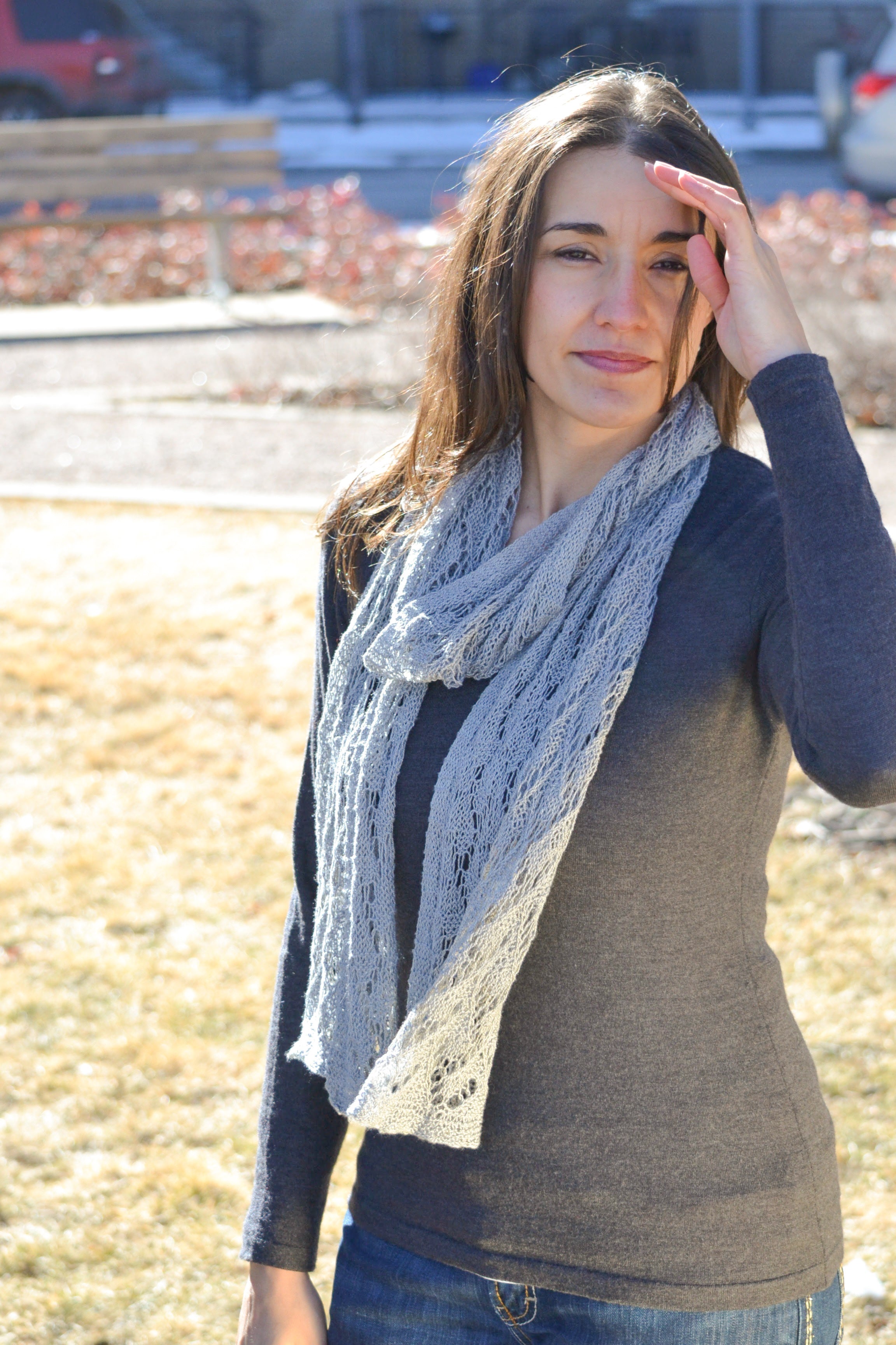 A woman with long brown hair wears the Shangri-La Knit Shawl, a lightweight lace wrap, over a dark sweater as she stands outside on a sunny day, shielding her eyes. Grass, benches, and a blurred red car appear in the background.