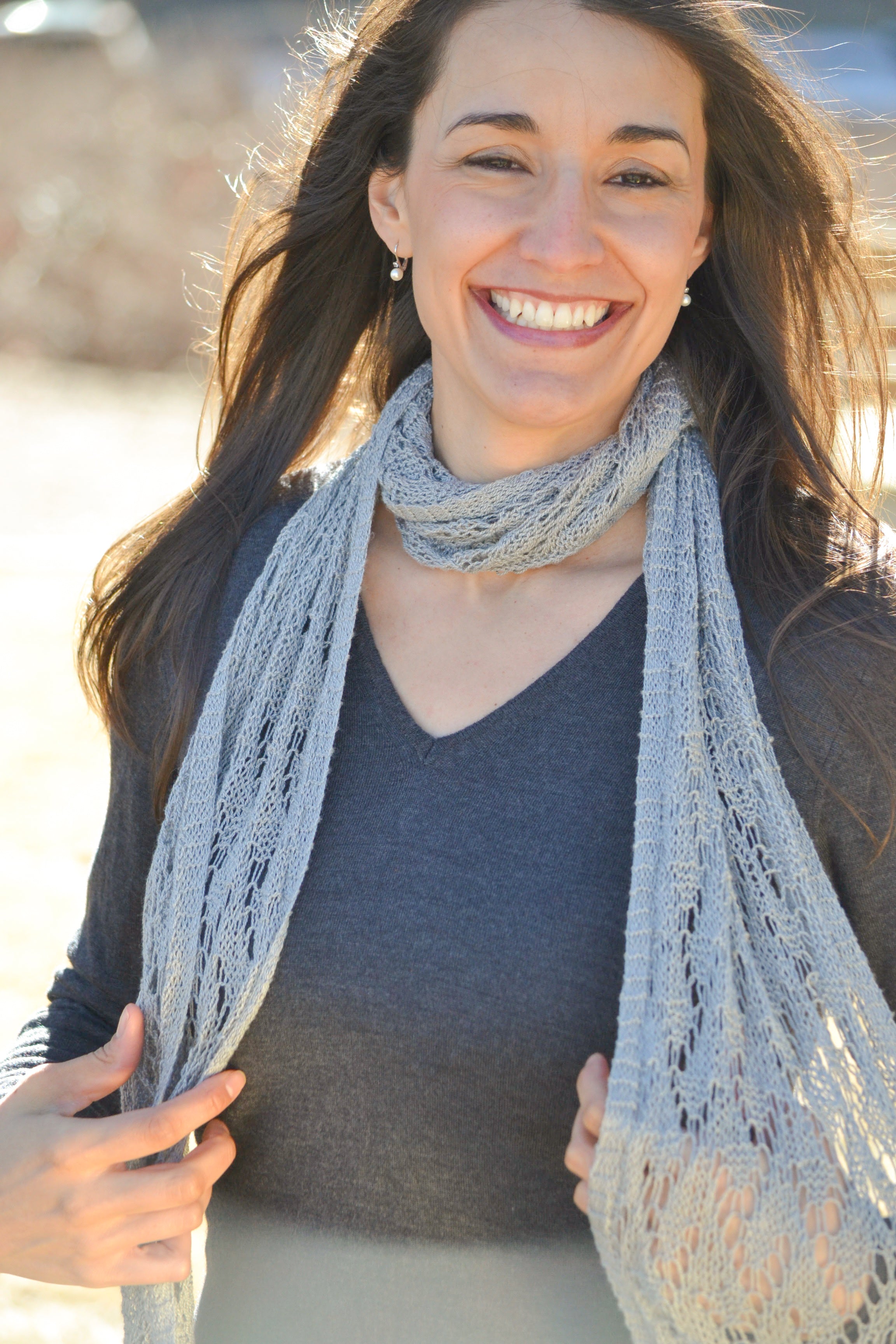 A woman with long brown hair smiles outdoors, wearing a dark gray V-neck shirt and a Shangri-La Knit Shawl—a lightweight lace shawl (scarf or wrap) from the PDF pattern. The softly blurred, sunlit background enhances the look.