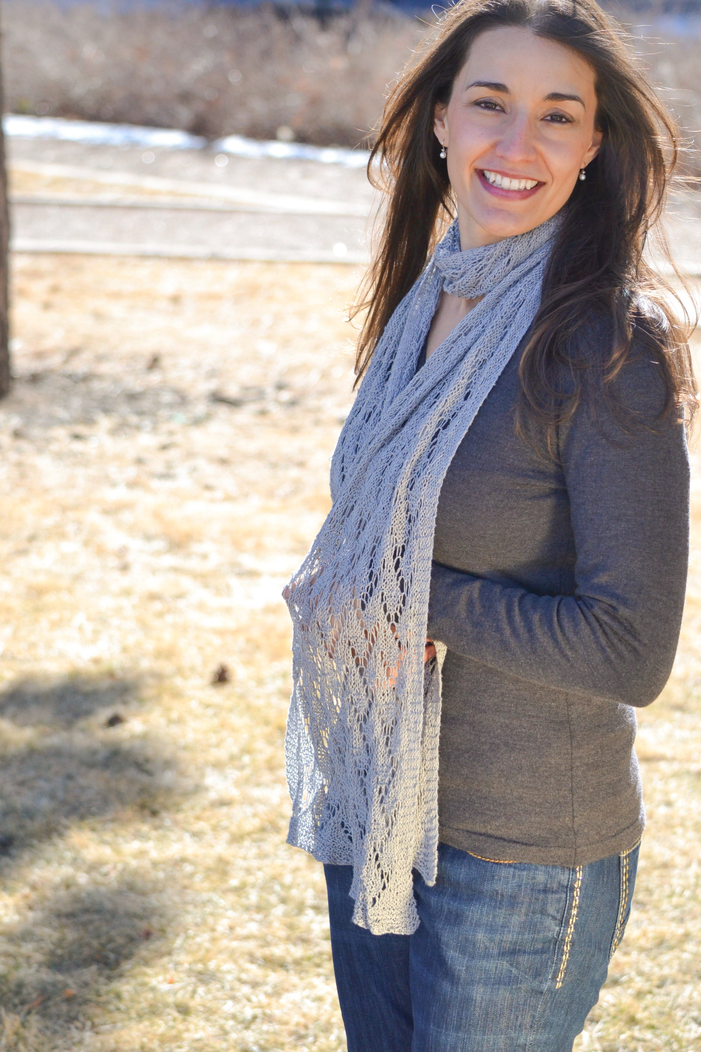 A woman with long brown hair smiles outdoors in the sun, wearing a gray long-sleeve shirt, blue jeans, and the Shangri-La Knit Shawl Pattern— a lightweight lace shawl (knitting PDF). Dry grass and trees are in the background.