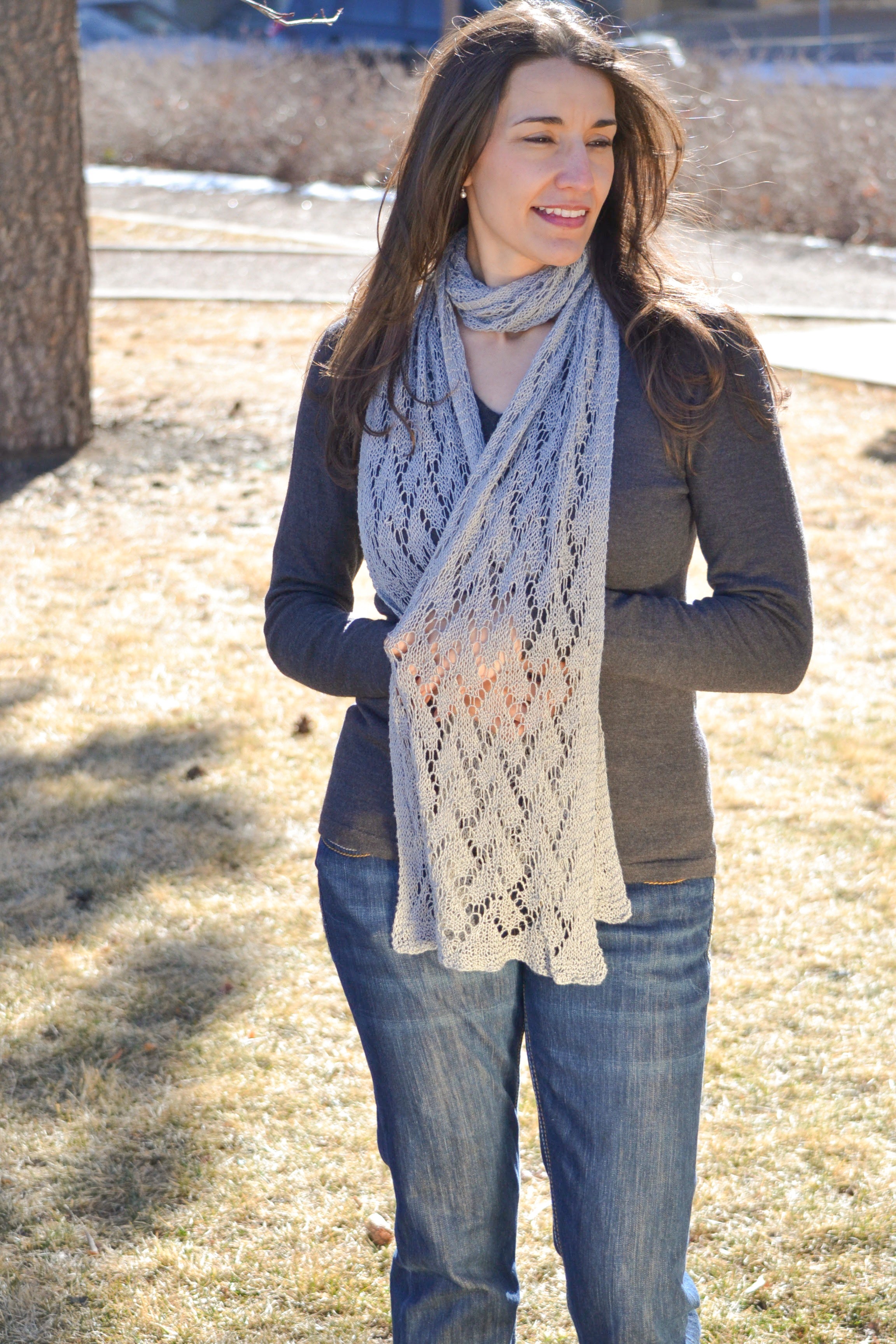 A woman with long brown hair smiles outdoors on a sunny day, wearing blue jeans, a gray long-sleeve top, and the Shangri-La Knit Shawl Pattern—a lightweight lace shawl (PDF pattern for scarf, shawl, or wrap) in light gray.