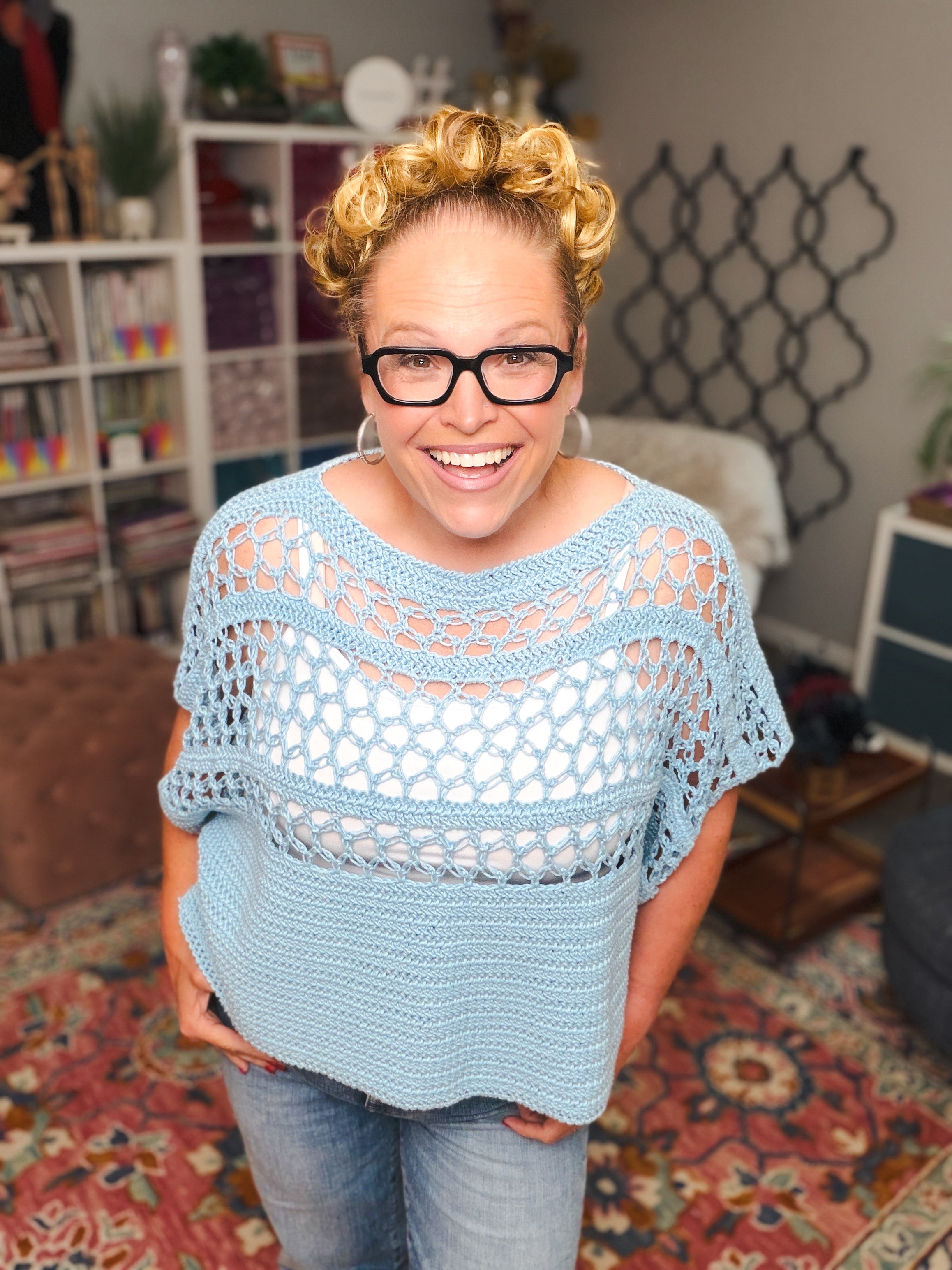 A woman with curly blond hair and glasses smiles indoors, wearing a light blue top made from the Solomon’s Knot Crochet Tee Pattern – a lightweight, size-inclusive summer crochet PDF. Behind her are colorful décor, bookshelves, and patterned rugs.