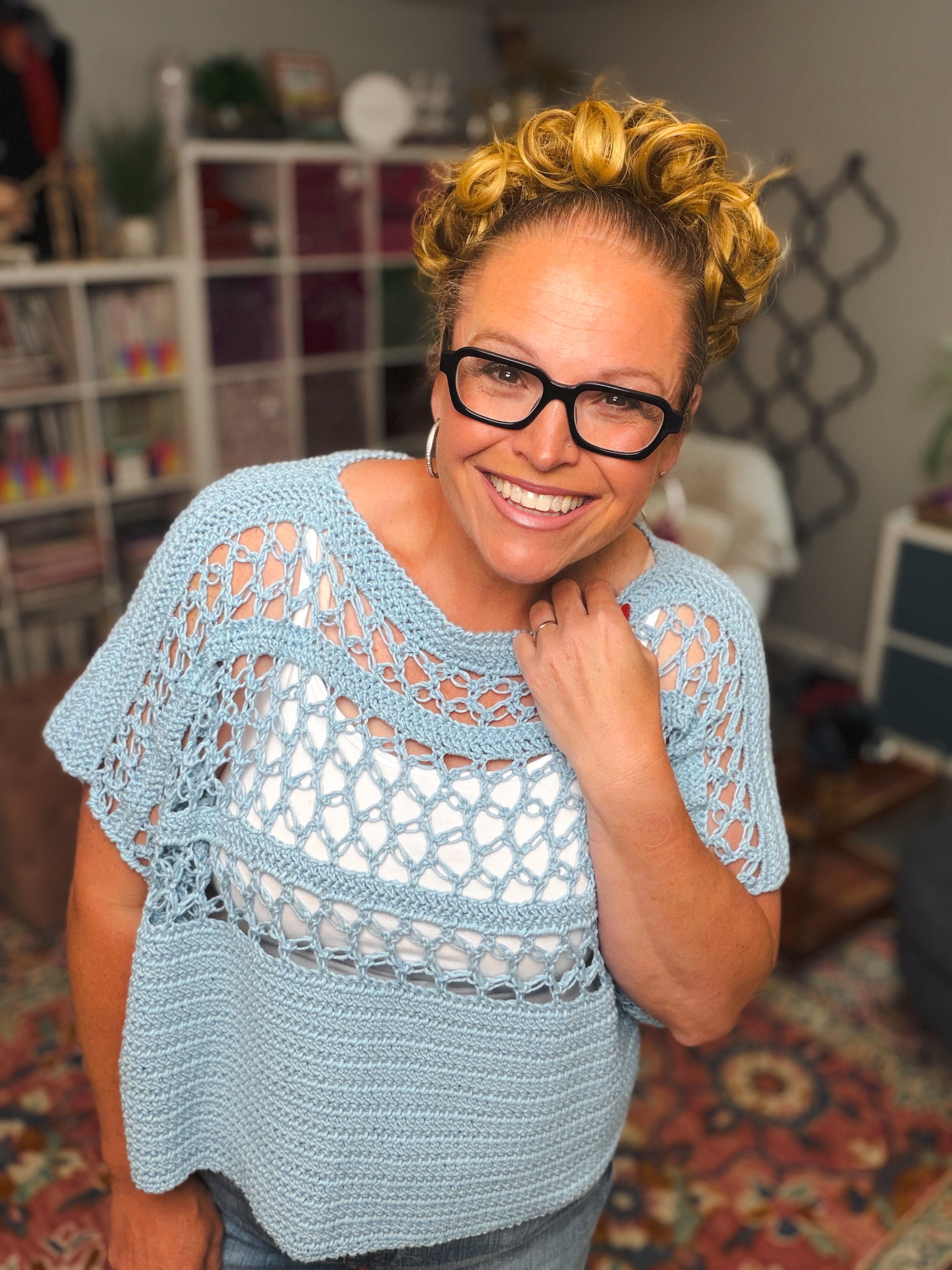 A smiling woman with curly blonde hair and glasses wears a light blue top made using the Solomon’s Knot Crochet Tee Pattern – Lightweight Summer Crochet Top (Size Inclusive) PDF, standing indoors with bookshelves and colorful decor behind her.