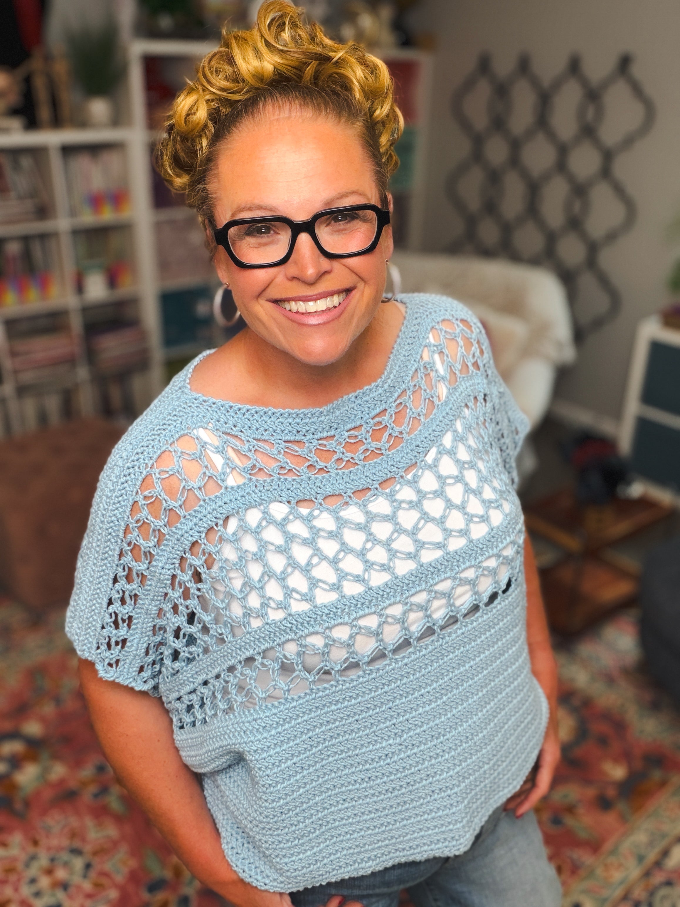 Smiling woman with short curly hair and glasses wears a light blue Solomon’s Knot Crochet Tee, made from the Lightweight Summer Crochet Top (Size Inclusive) PDF pattern, standing indoors with bookshelves and colorful decor in the background.