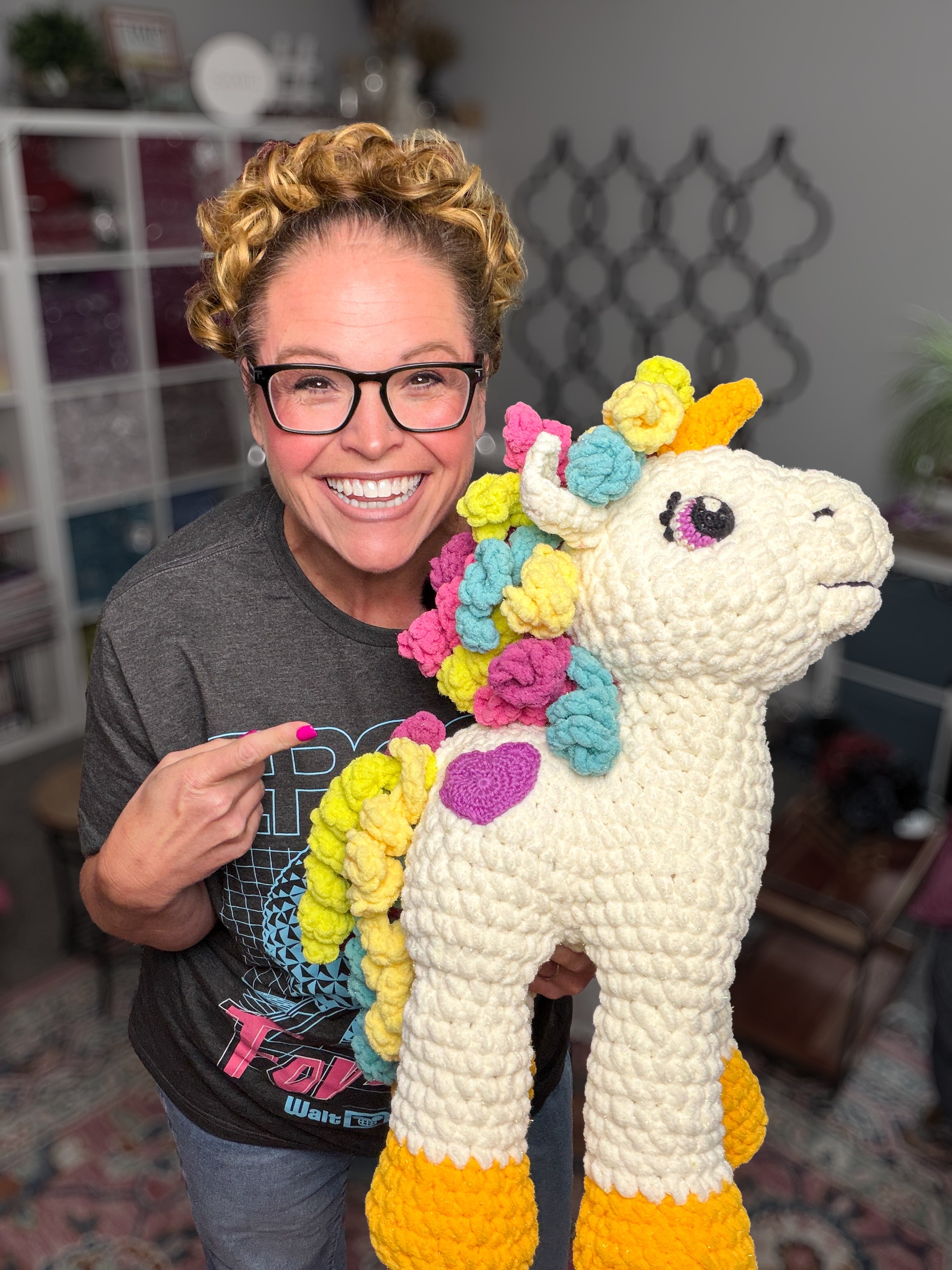 A smiling woman with curly hair and glasses points to a large, colorful unicorn with a rainbow mane—an adorable creation made from the Sparkle Crochet Unicorn Amigurumi Pattern—in a cozy, yarn-filled room.