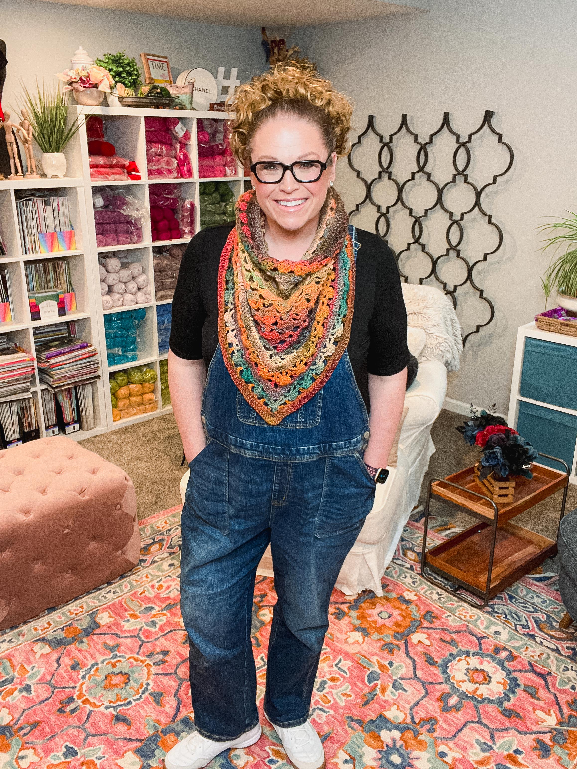 Wearing denim overalls over a black shirt and white shoes, a person with curly hair and glasses stands smiling in a cozy, colorful room filled with yarn, books, and plants. Around their neck is a vibrant bandana cowl—crocheted in the delicate lacy shell stitch using the Sunday Chill Crochet Bandana Cowl Pattern.