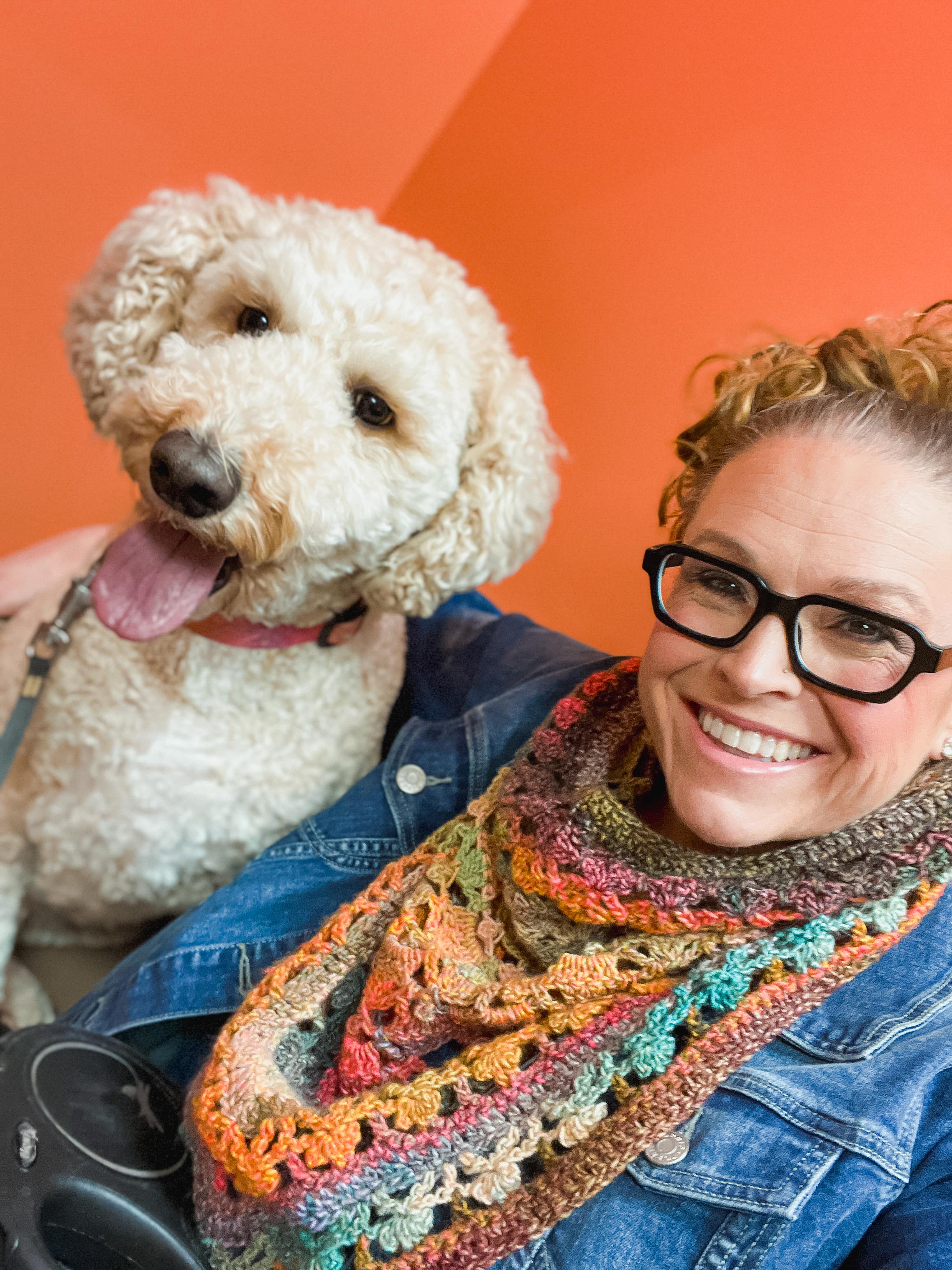 A smiling woman with glasses and curly hair poses with a happy, fluffy tan dog against an orange background. She wears a denim jacket and the Sunday Chill Crochet Bandana Cowl, featuring colorful, lacy shell stitching in a triangular shape. The dog sports a pink collar.