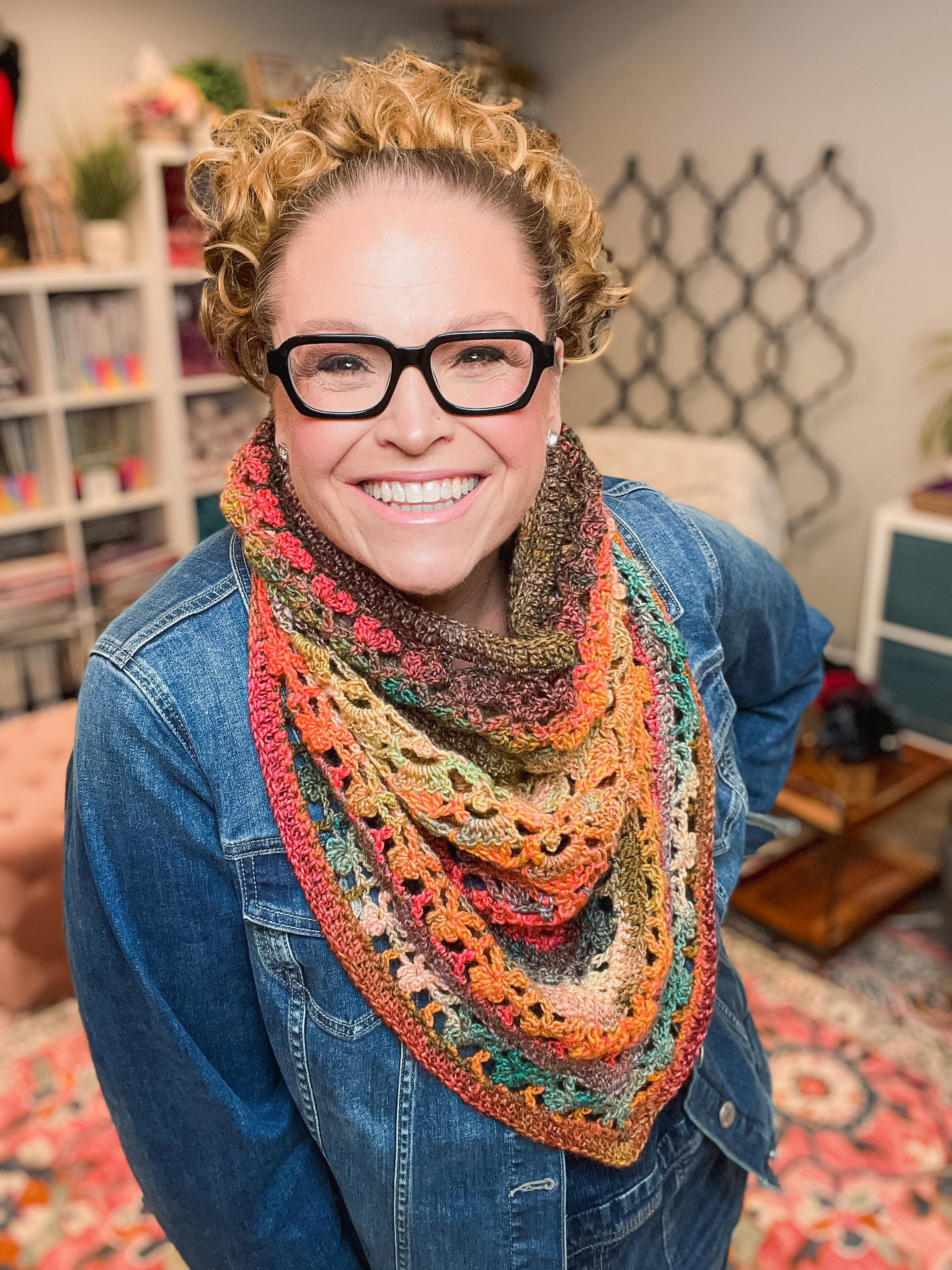 A smiling person with curly hair and glasses stands in a cozy room filled with shelves, yarn, and patterned decor, wearing a blue denim jacket and the Sunday Chill Crochet Bandana Cowl Pattern—a colorful, triangular crochet shawl.