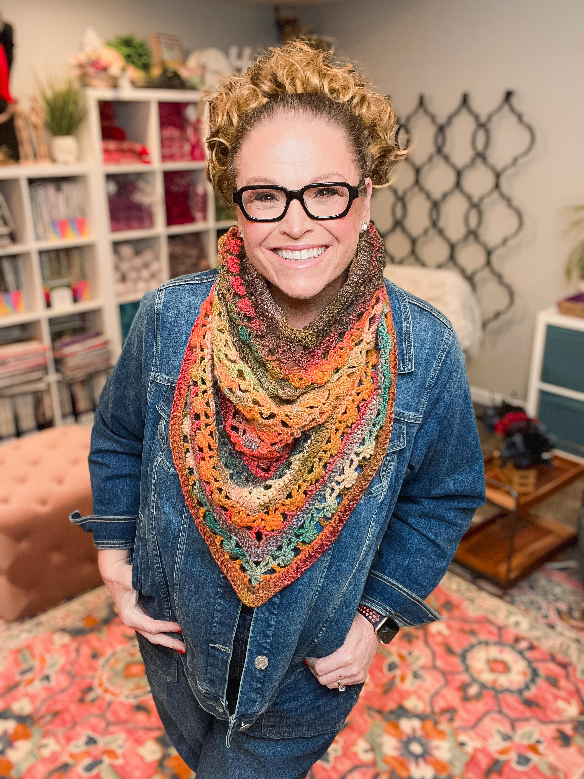 A smiling person with curly hair and glasses wears the Sunday Chill Crochet Bandana Cowl over a denim jacket, standing in a cozy, colorful room filled with bookshelves and yarn.