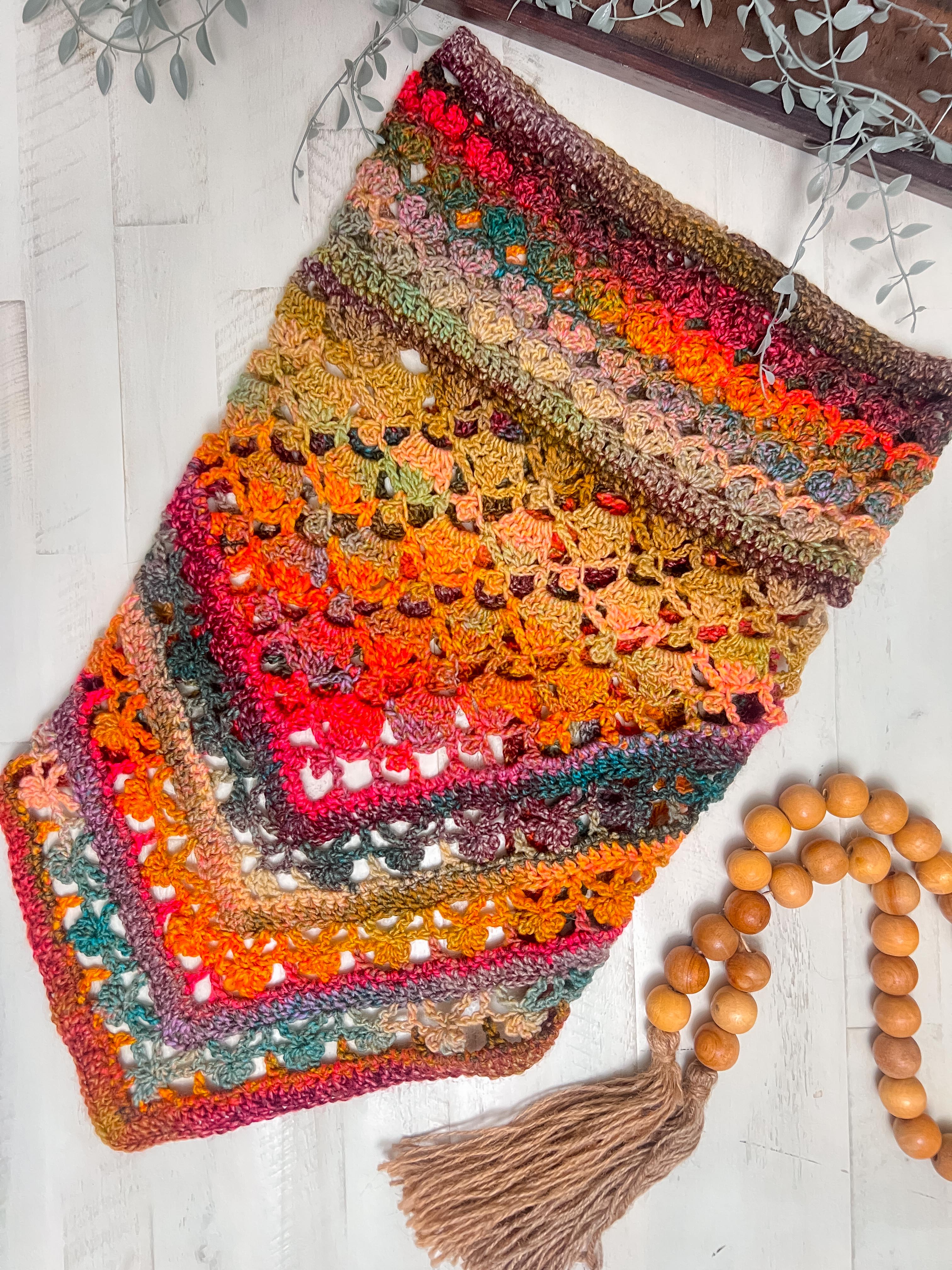 The Sunday Chill Crochet Bandana Cowl, featuring intricate lacy shell stitch patterns in shades of orange, yellow, green, and red, is displayed on a white wooden surface beside wooden beads with a tassel and green foliage.