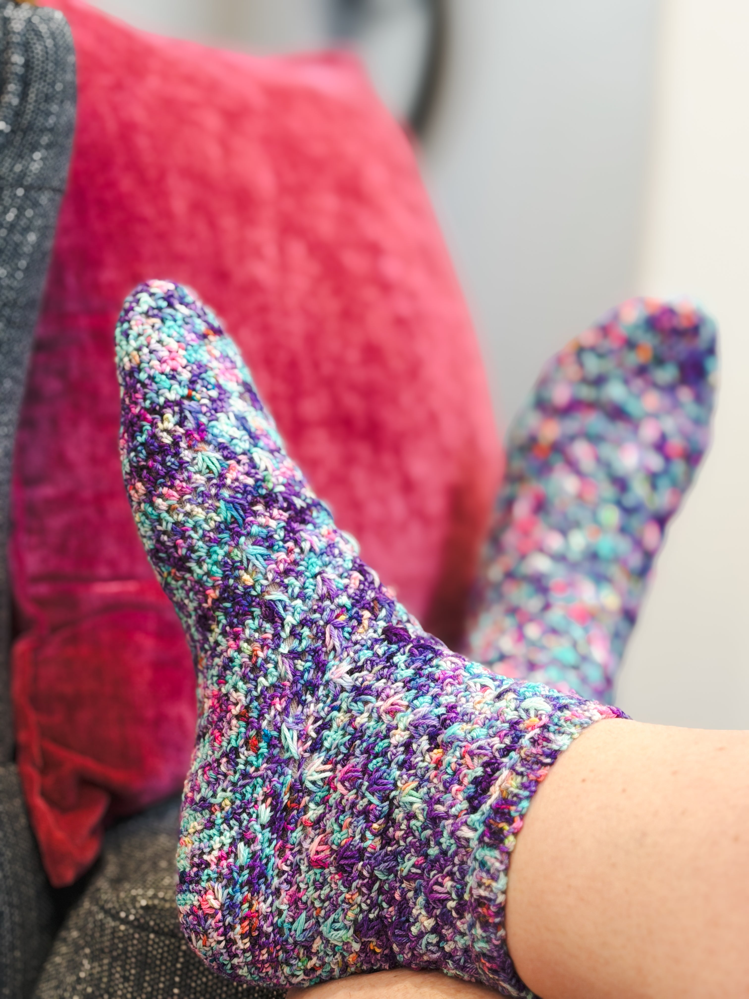 Close-up of a person’s feet wearing colorful, handmade socks created using the Sweet Nothings Crochet Socks Pattern, resting on a sofa with a bright pink pillow in the background.