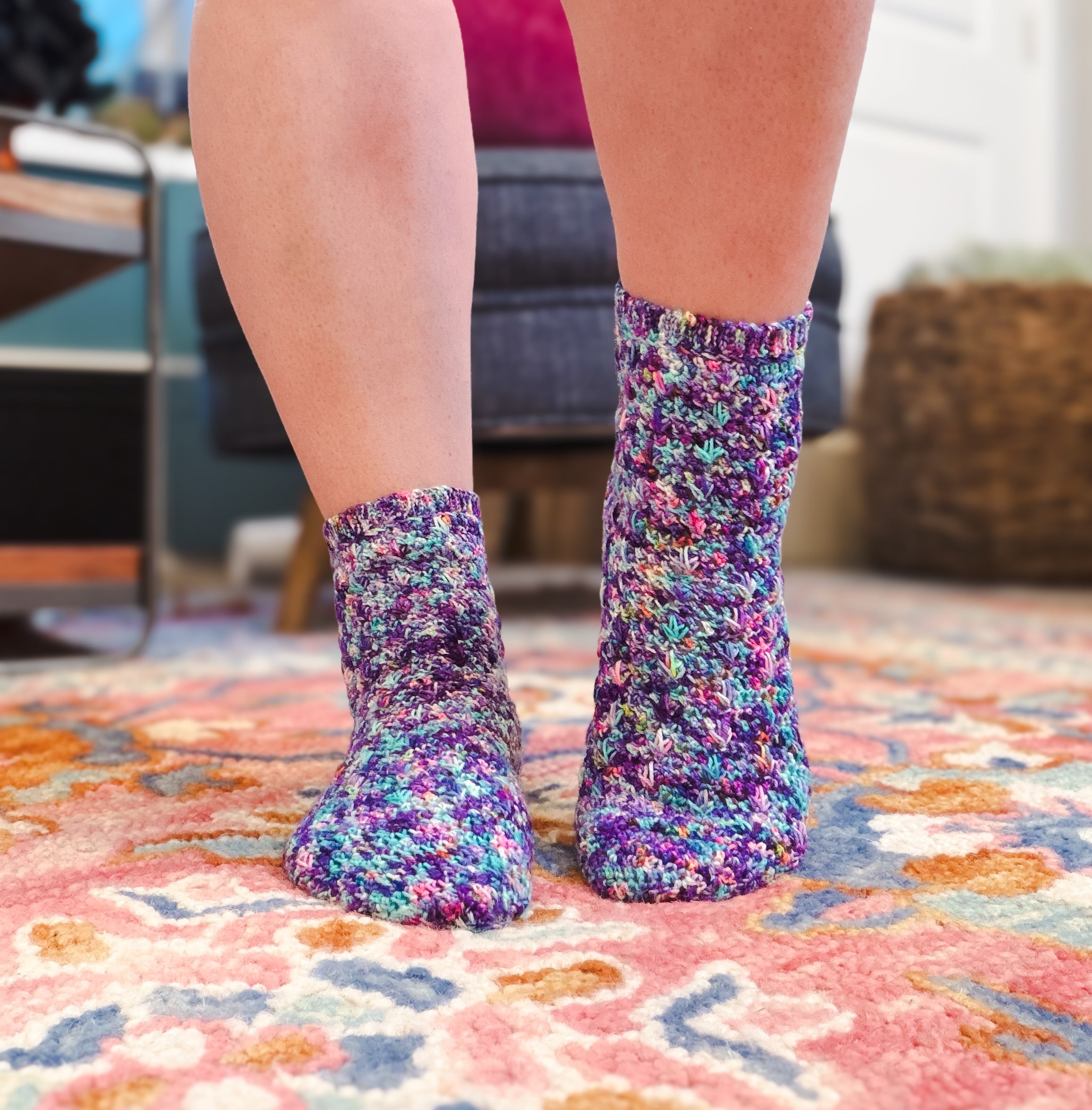 Two feet wearing colorful, speckled knit socks stand on a multicolored patterned rug in a cozy indoor setting, showcasing the unique design of the Sweet Nothings Crochet Socks Pattern and the intricate texture reminiscent of the textured spike stitch.