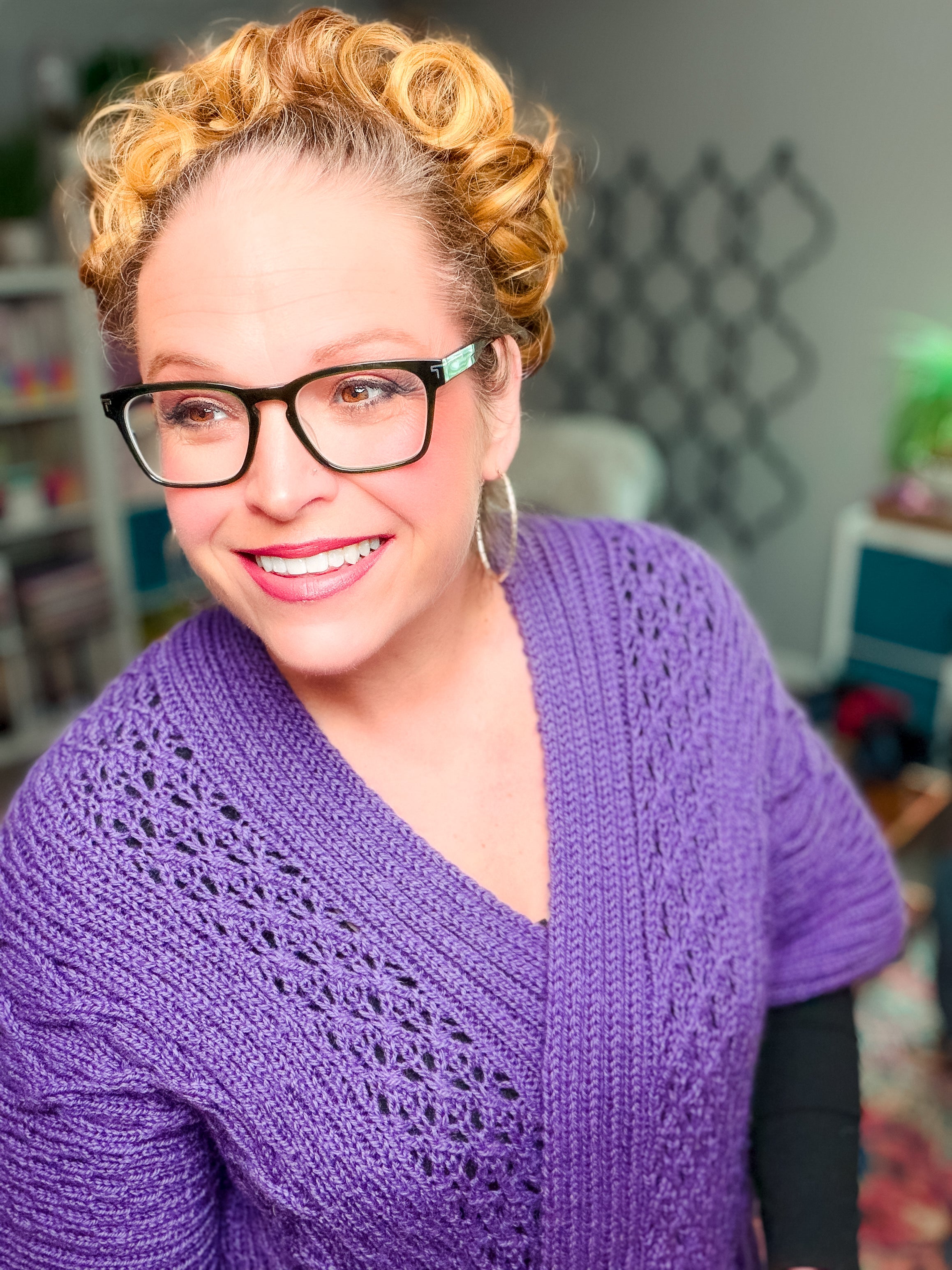 Smiling woman with curly blonde hair, black glasses, and hoop earrings models the Sycamore Knit Ruana Pattern—a stylish purple worsted weight wool cardigan worn over black sleeves—standing indoors against a blurred background. This project is perfect inspiration for intermediate knitters ready to take on their next ruana pattern.