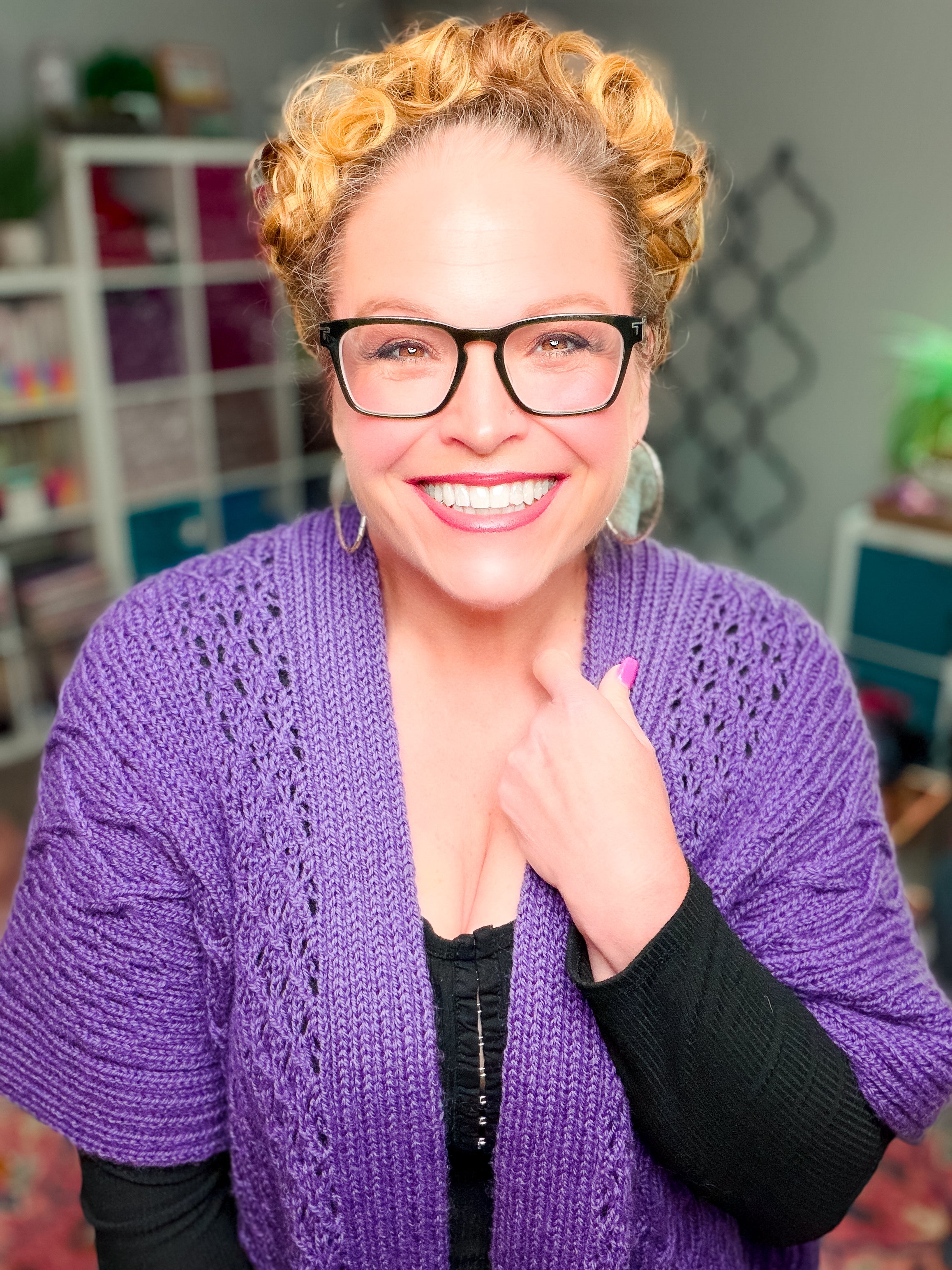 A smiling person with curly blonde hair and glasses wears the Sycamore Knit Ruana—a textured wrap in rich purple worsted weight wool—over a black top, standing in a bright room with shelves and colorful decor in the background.