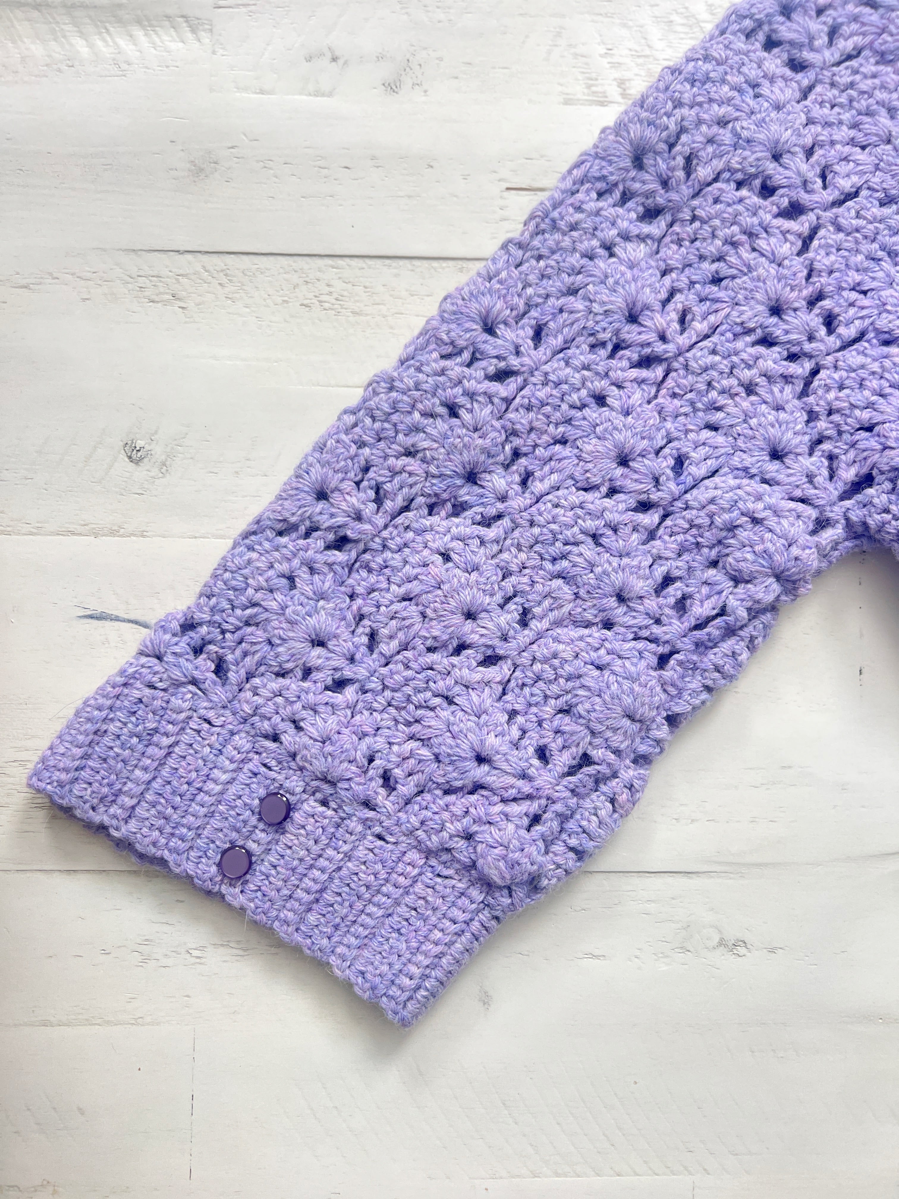 A close-up of a lavender crocheted sleeve with a textured floral pattern and ribbed cuff, featuring two small purple buttons on the cuff, laid on a white wooden surface—perfectly showcasing the delicate details of the Tatiana Crochet Lace Tunic – Lace Crochet Top PDF (Sizes S–2X) for a beautiful, lightweight crochet tunic.