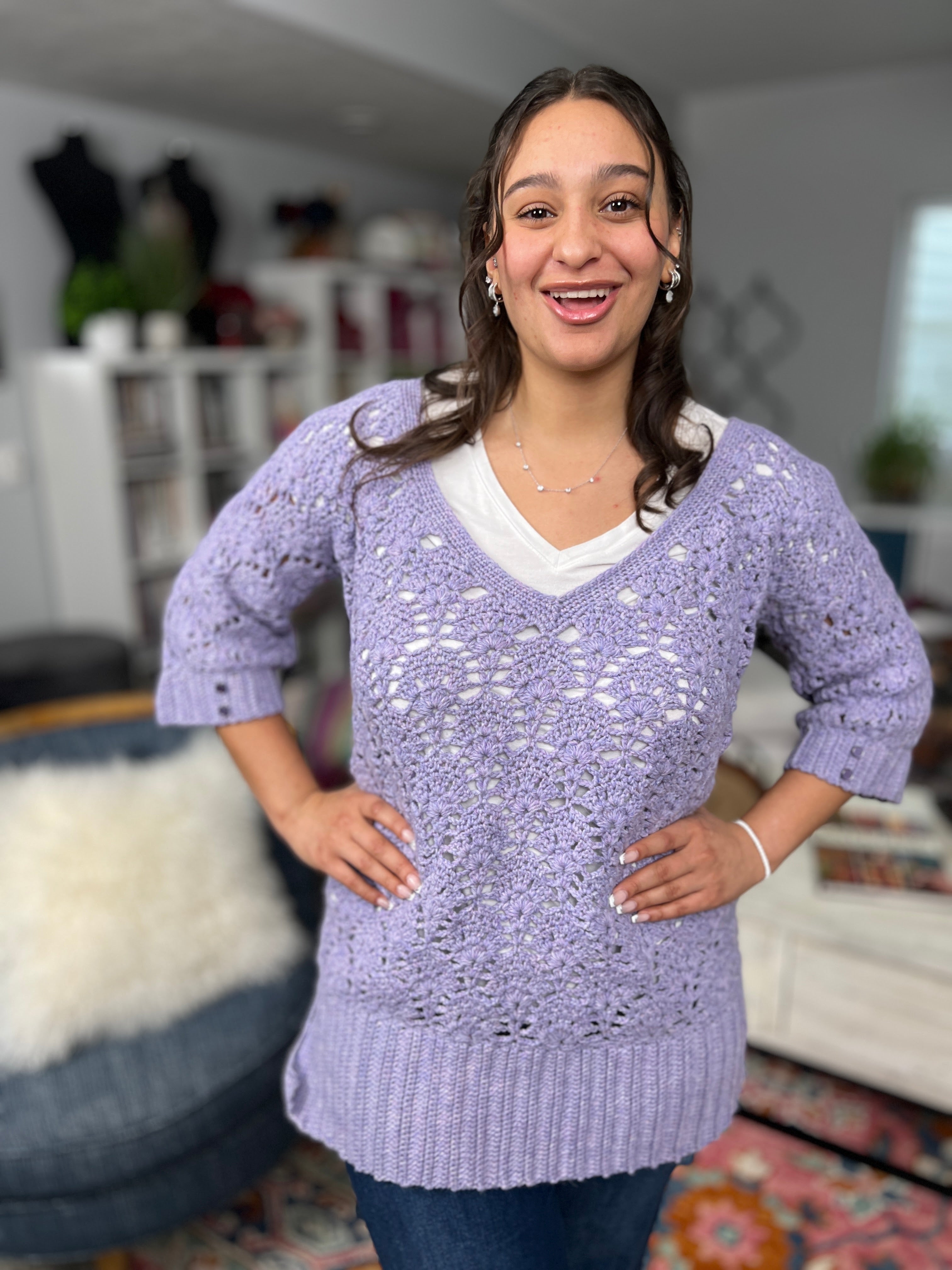 A woman smiles with her hands on her hips, wearing the Tatiana Crochet Lace Tunic—a light purple lace crochet top (pattern available in sizes S–2X)—styled over a white shirt. She stands in a cozy, well-decorated living room featuring bookshelves and textured decor in the background.