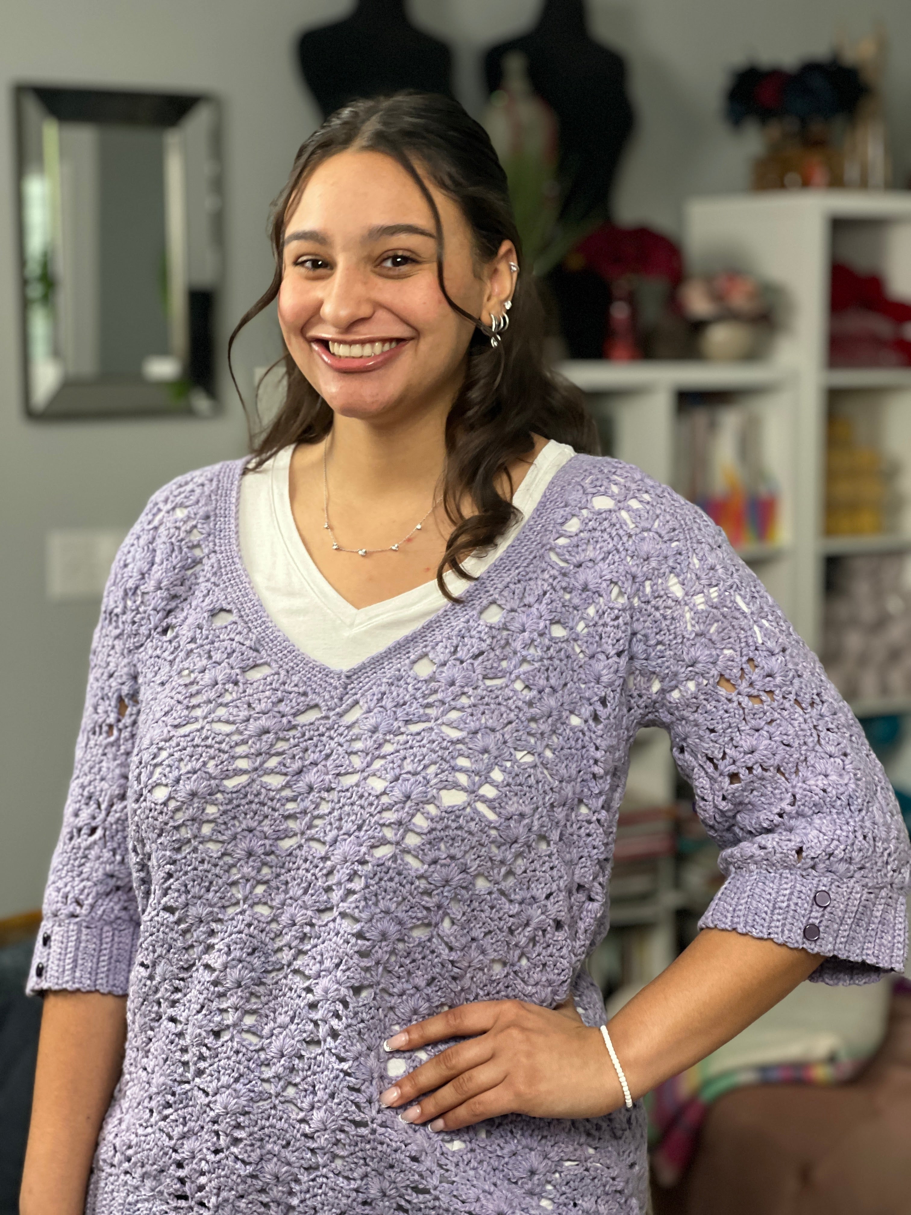 A woman with long dark hair, wearing a white shirt layered under the Tatiana Crochet Lace Tunic—a delicate lavender lace crochet top—smiles while standing indoors in a cozy room with bookshelves and mannequins in the background.