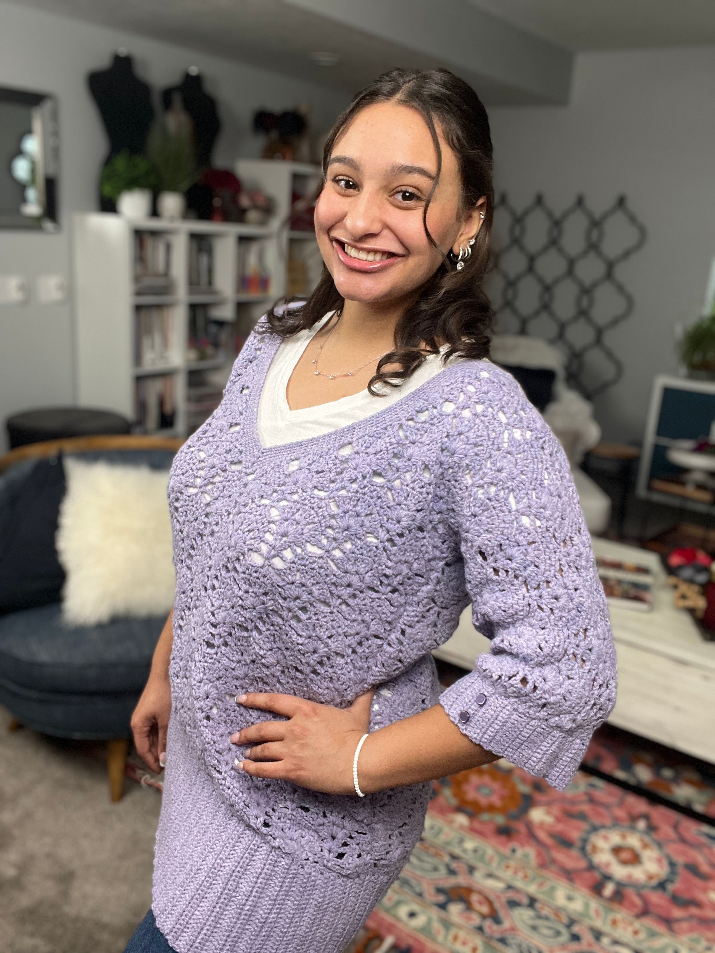Wearing the Tatiana Crochet Lace Tunic—a delicate lavender V-neck crochet top—a woman smiles as she stands in a cozy living room adorned with bookshelves, plants, and vibrant rugs.