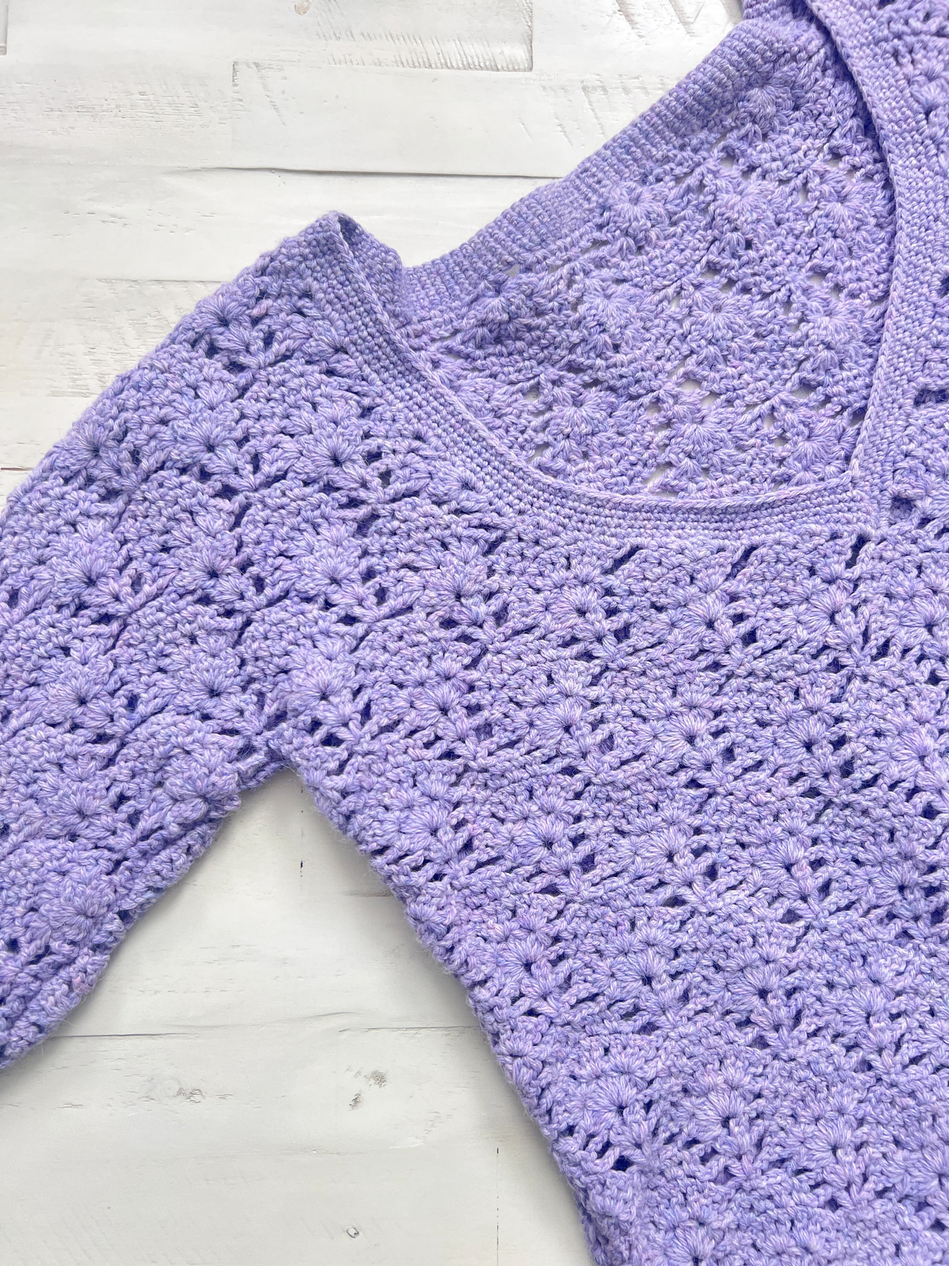 A close-up of the Tatiana Crochet Lace Tunic, a lavender lace crochet top featuring a scalloped floral pattern and a V-shaped neckline, laid flat on a white wooden surface. This lightweight tunic is crafted using the Lace Crochet Top PDF (Sizes S–2X) for an elegant finish.