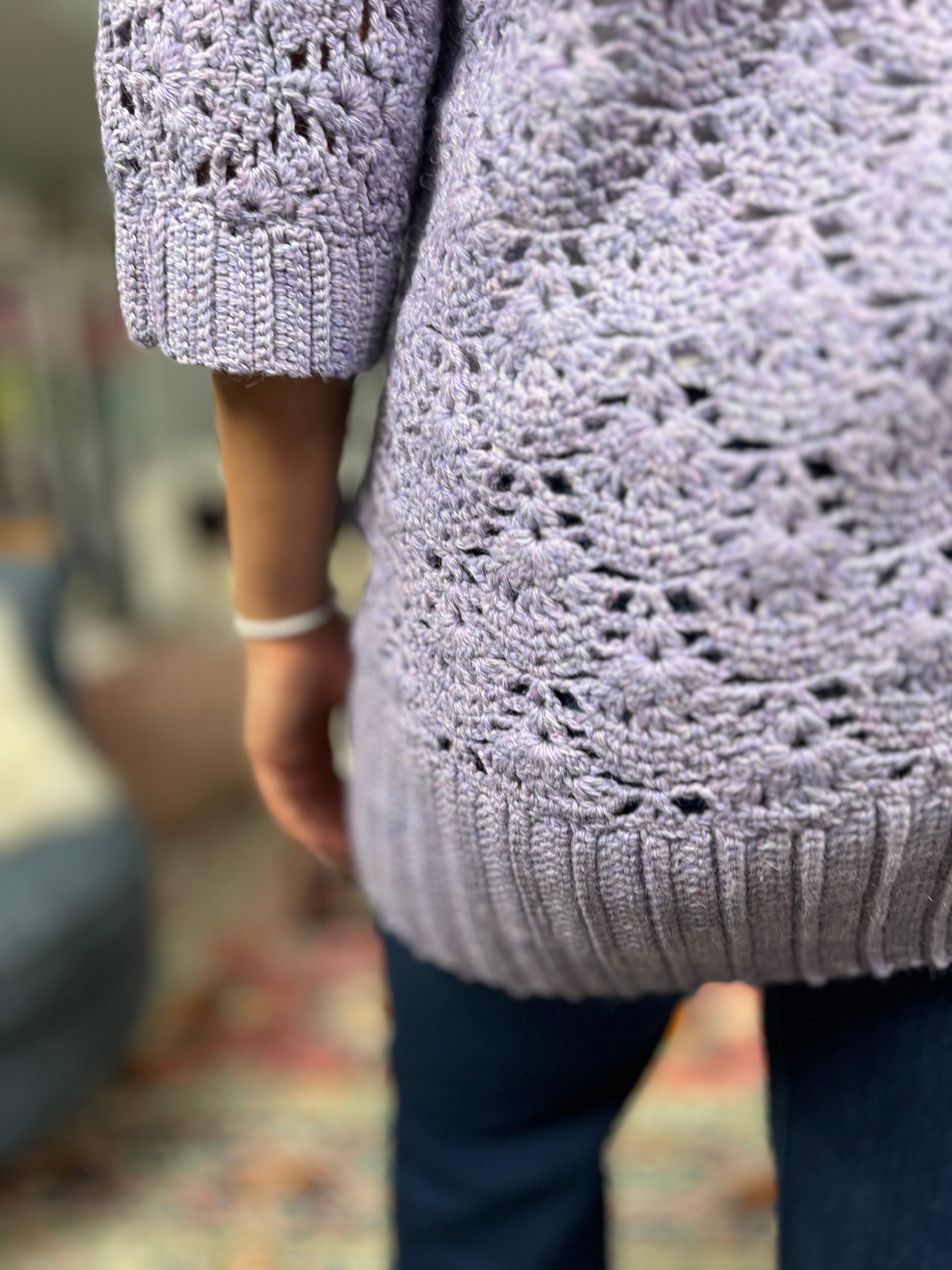 A person wearing the Tatiana Crochet Lace Tunic – Lace Crochet Top (PDF, Sizes S–2X) in light purple and dark pants stands indoors, shown from the back and slightly to the side. The background is softly blurred.