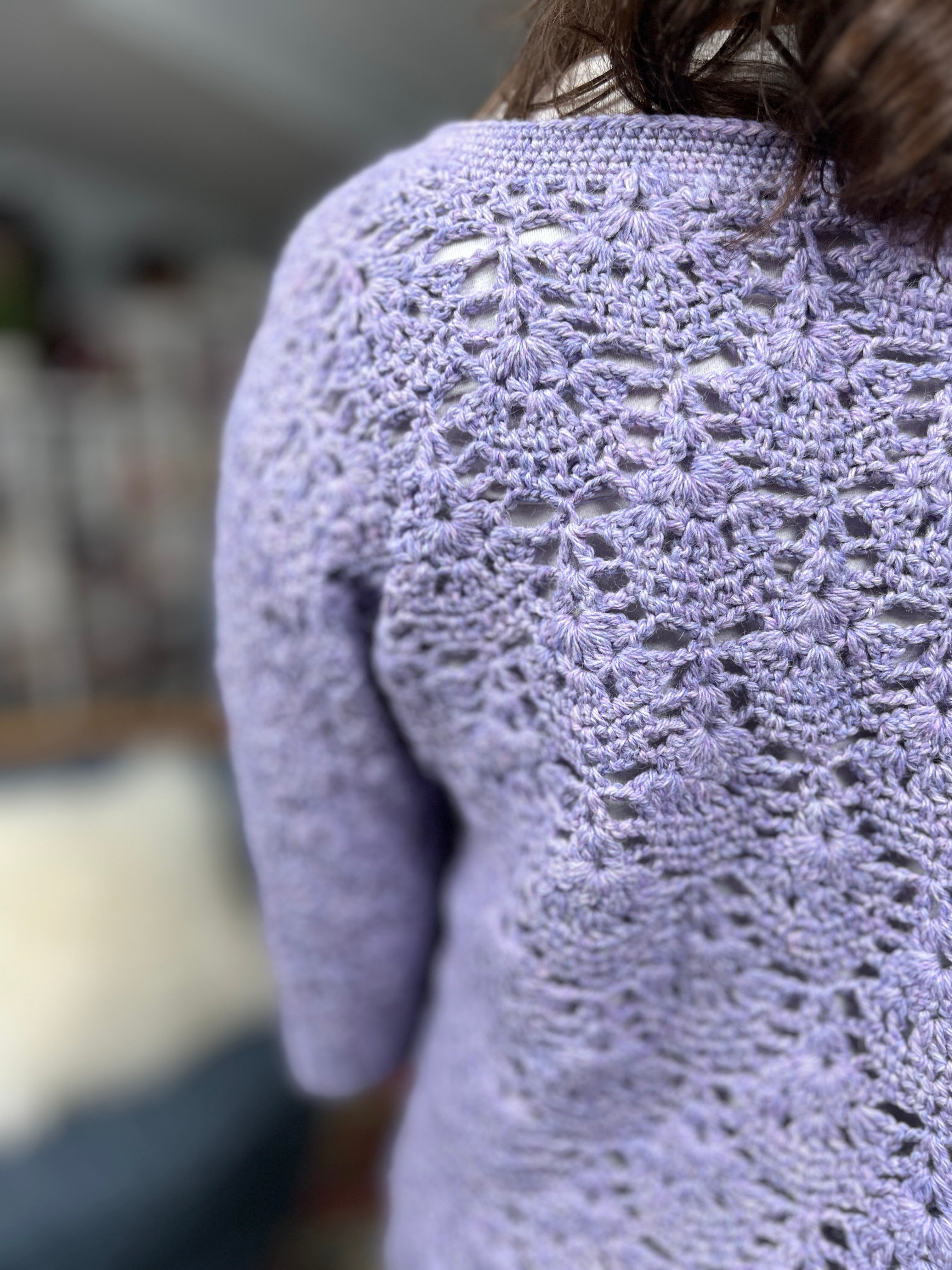 A person is wearing the Tatiana Crochet Lace Tunic—a light purple, lightweight top featuring a delicate floral lace crochet pattern. The focus is on the back and sleeve, highlighting the detailed lace stitch crochet work of this tunic. The background is blurred.