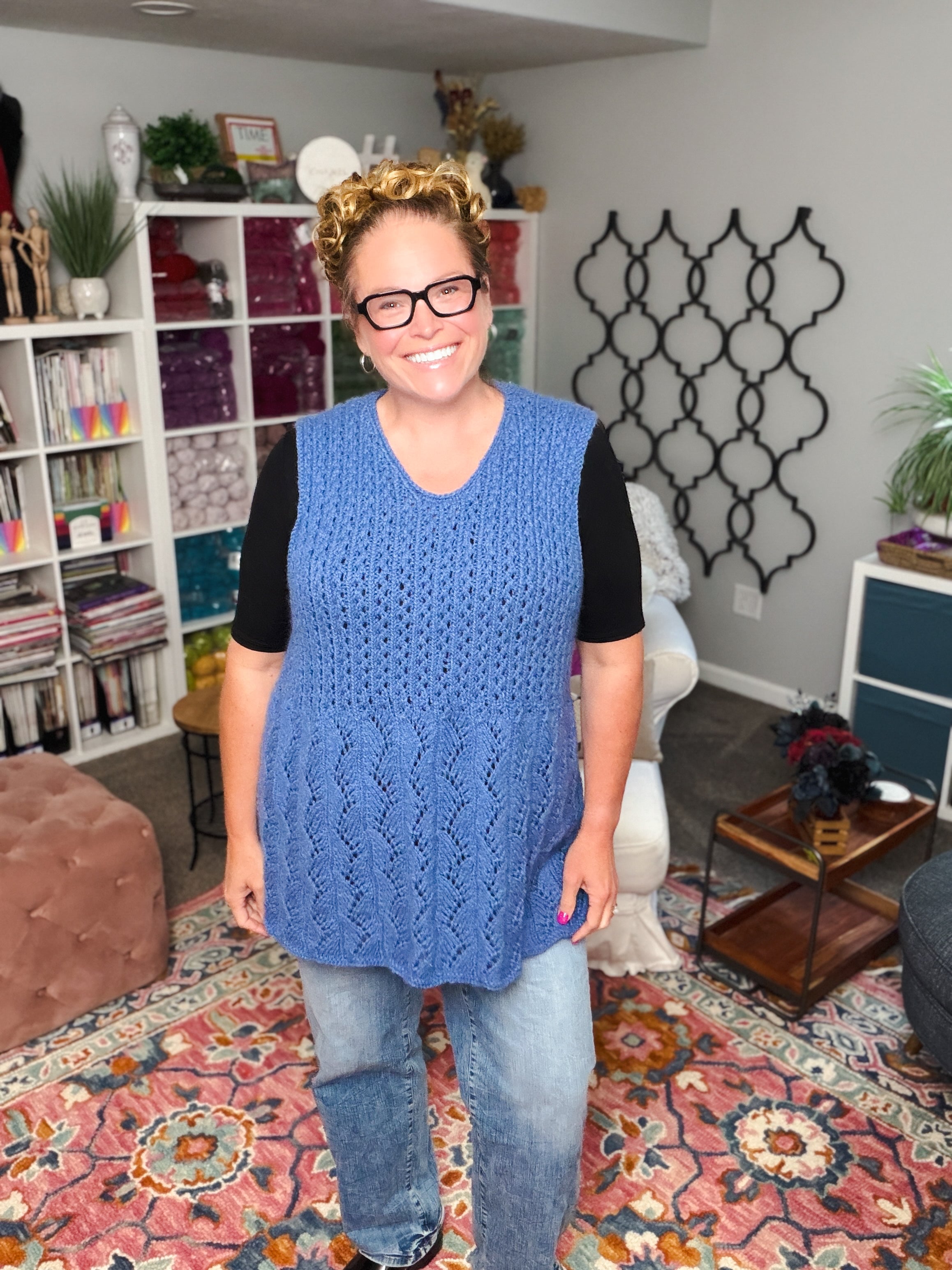 A smiling woman with glasses and curly hair models the Thistlevine Knit Tunic Pattern—a lace knit sleeveless sweater (XS–5X)—over a black shirt and jeans, standing in a cozy room with a colorful rug, yarn shelves, and decor.