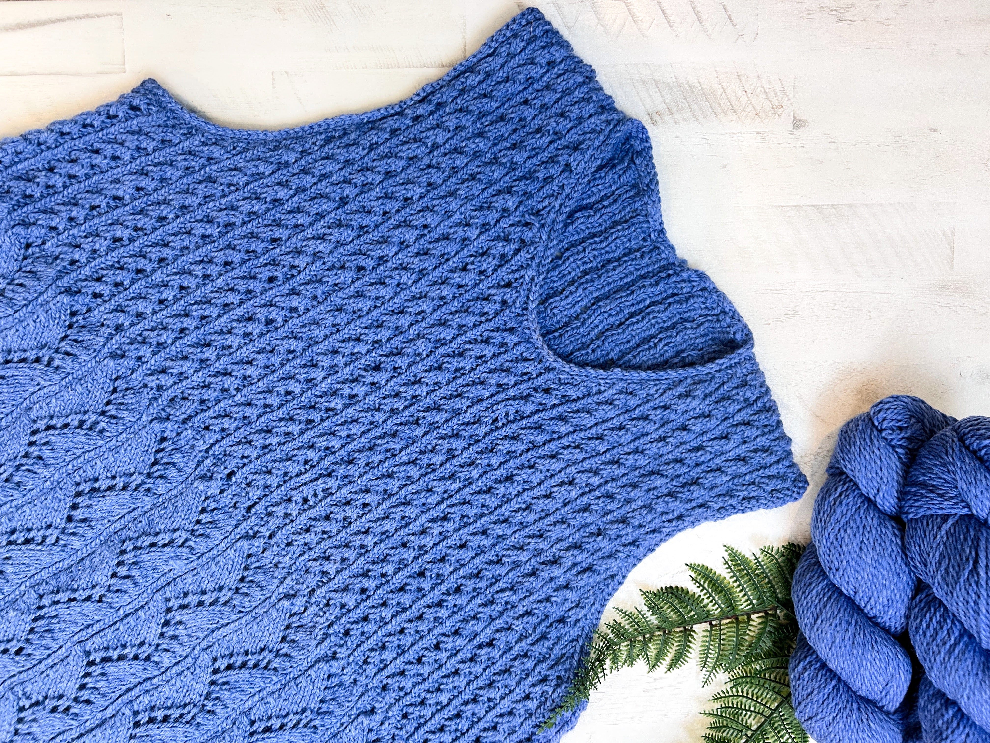 A close-up of the Thistlevine Knit Tunic Pattern—a lace knit sleeveless sweater (PDF, XS–5X)—shown in blue on a light wood surface, with a matching blue yarn skein and fern leaf.