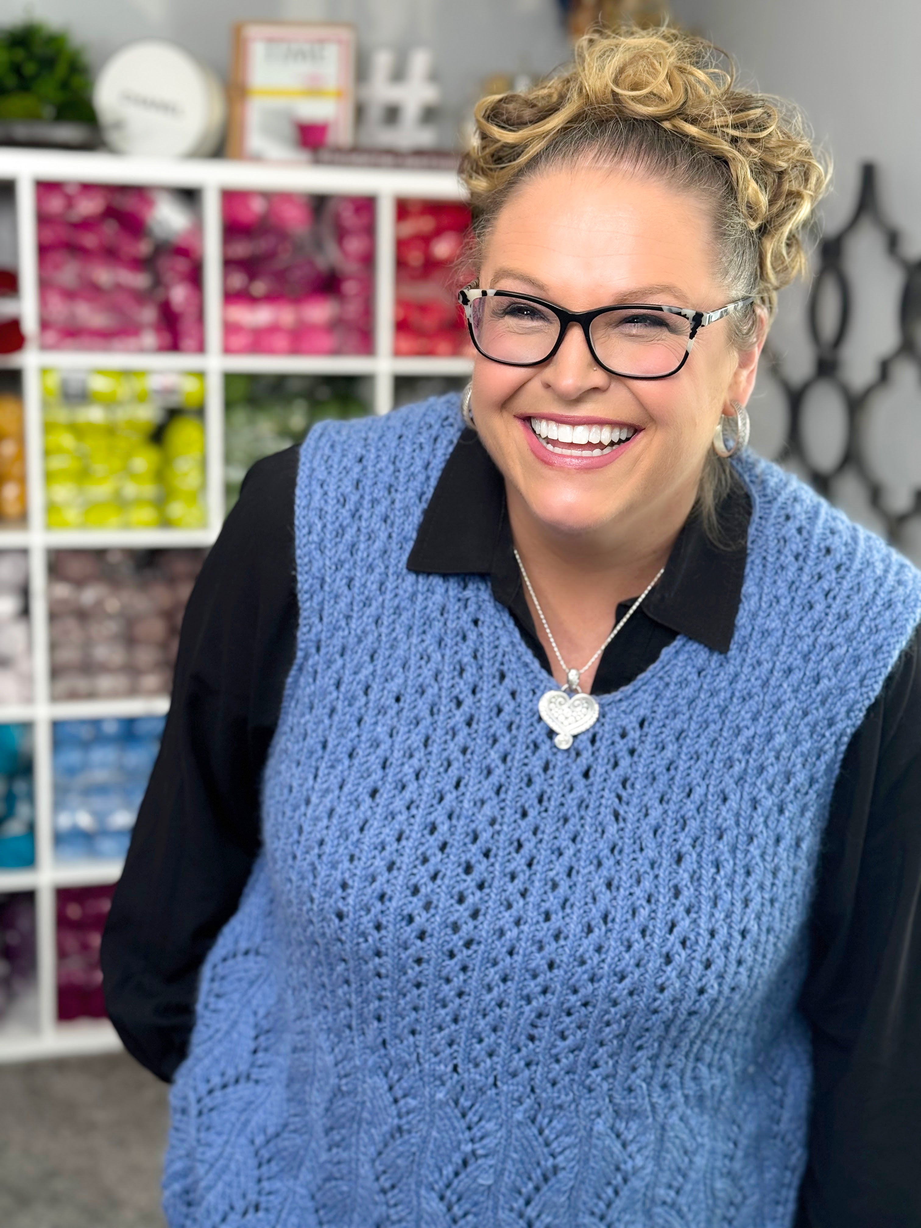 A smiling woman with curly hair and glasses wears the Thistlevine Knit Tunic—a lace knit sleeveless sweater (PDF pattern, XS–5X)—over a black shirt, standing before shelves of colorful yarn for a lively craft room vibe.