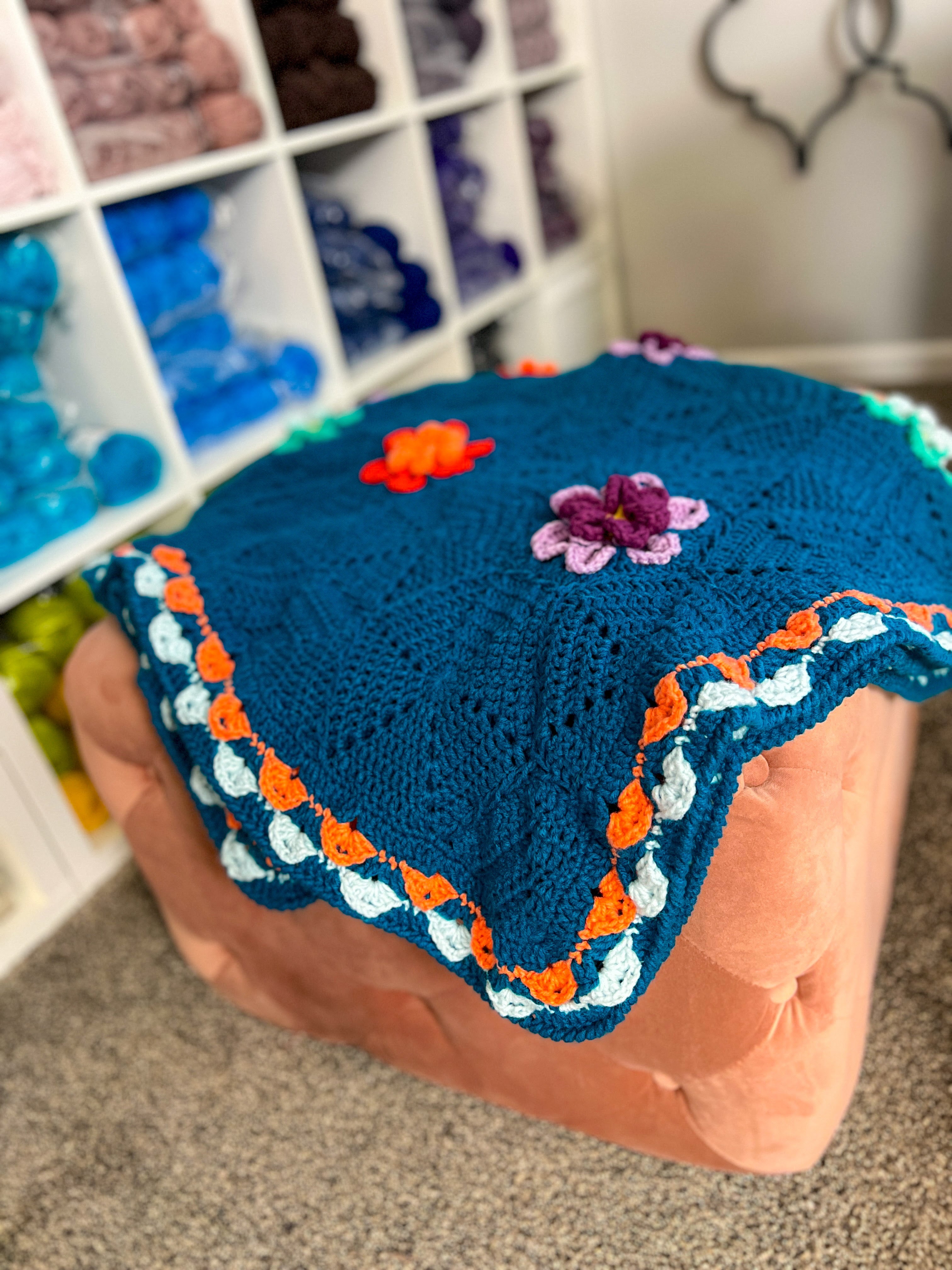 The Wildflower Hexagon Motif Crochet Blanket Pattern is showcased here in blue, adorned with vibrant floral accents and a delicate scalloped edge. Draped elegantly over a peach ottoman, with shelves of yarn in the background, this blanket highlights the timeless beauty of hexagon crochet motif patterns.