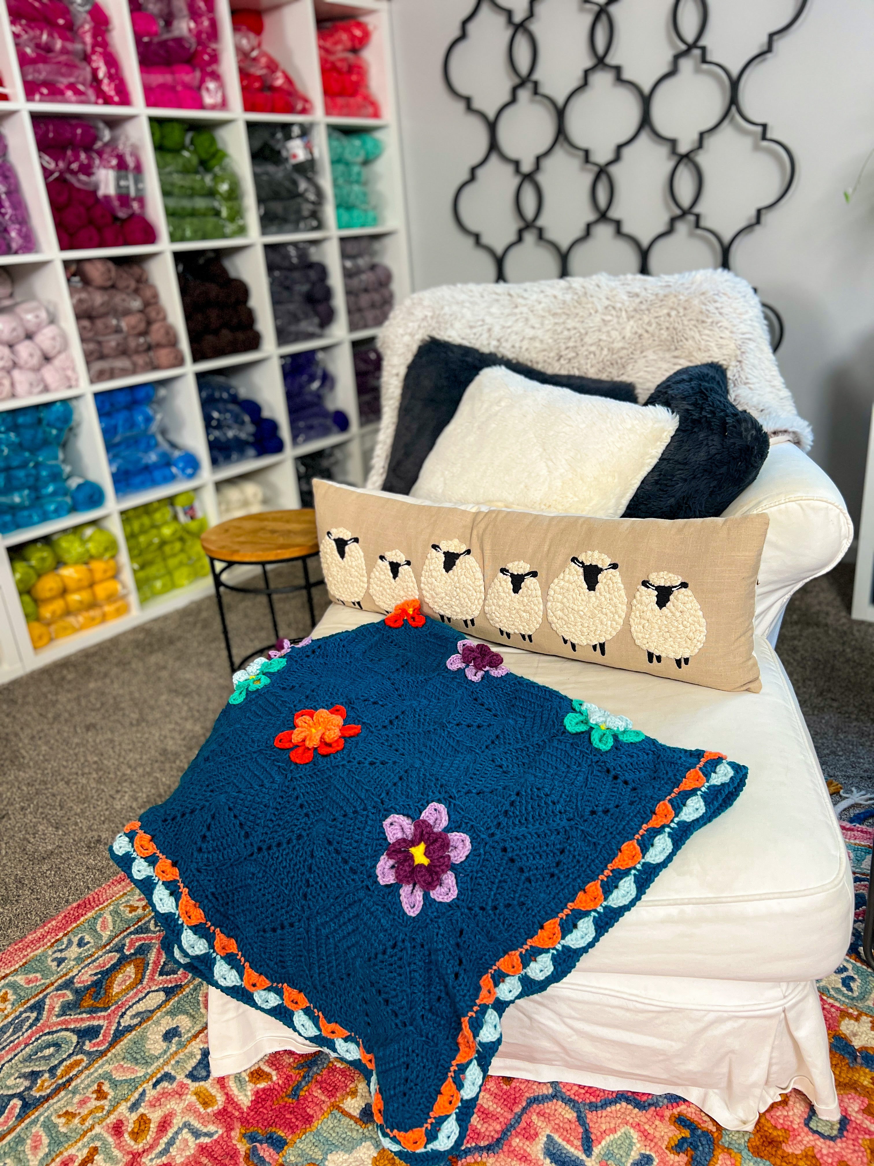 A cozy white chair with a sheep-themed pillow and the vibrant Wildflower Hexagon Motif Crochet Blanket draped over it sits beside a wall lined with organized yarn cubbies in assorted shades, above a bright, patterned rug on the floor.