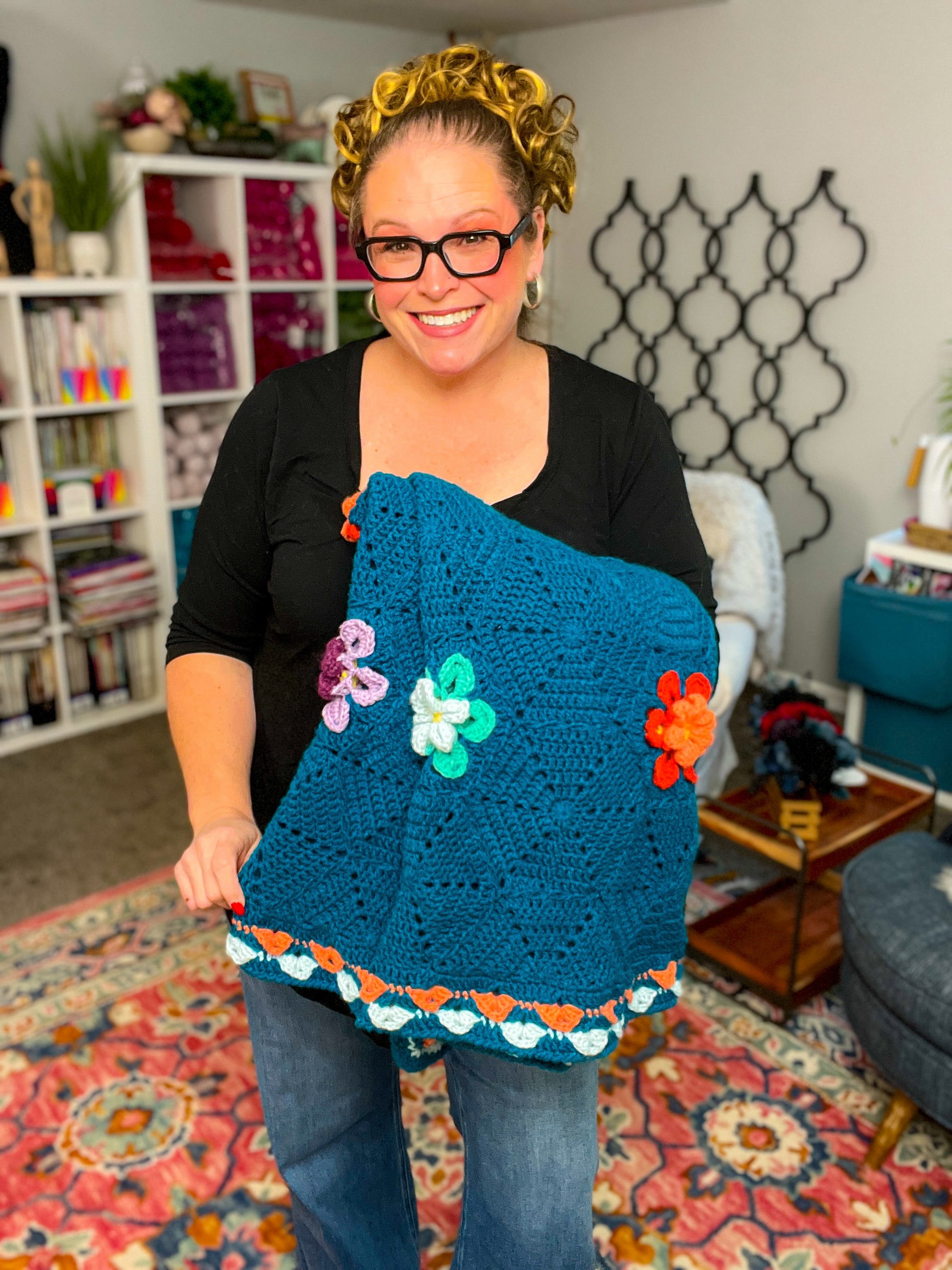 A smiling woman with glasses holds a blue blanket made from the Wildflower Hexagon Motif Crochet Blanket Pattern, beautifully adorned with colorful floral motifs, standing in a cozy, well-lit room filled with shelves, books, and yarn in the background.