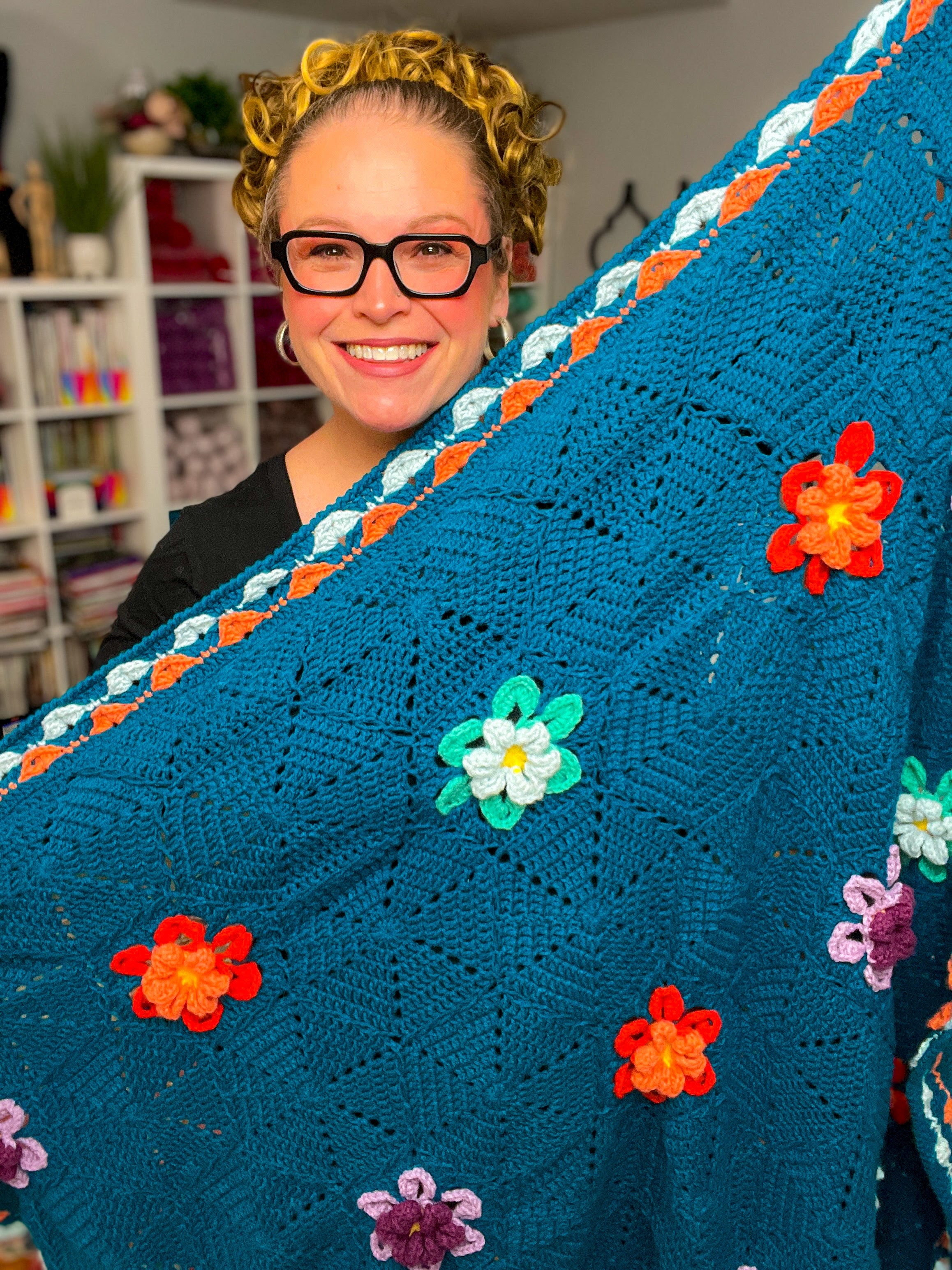 A smiling person with glasses holds up a cozy Wildflower Hexagon Motif Crochet Blanket, featuring blue tones and colorful 3D flower designs, in a creative workspace.