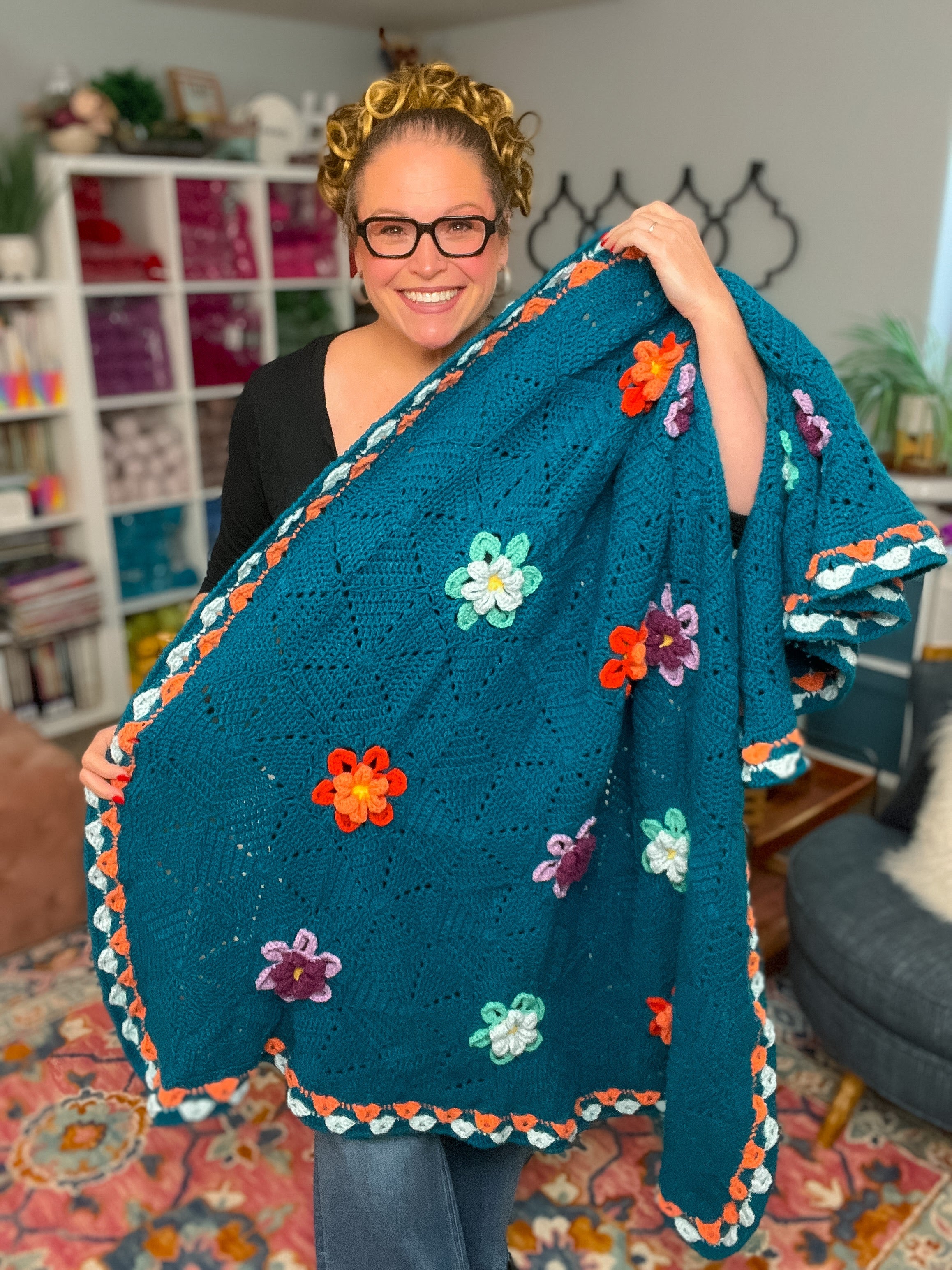 A smiling person with glasses holds up a teal Wildflower Hexagon Motif Crochet Blanket, decorated with colorful flower motifs, while standing in a cozy, yarn-filled craft room.