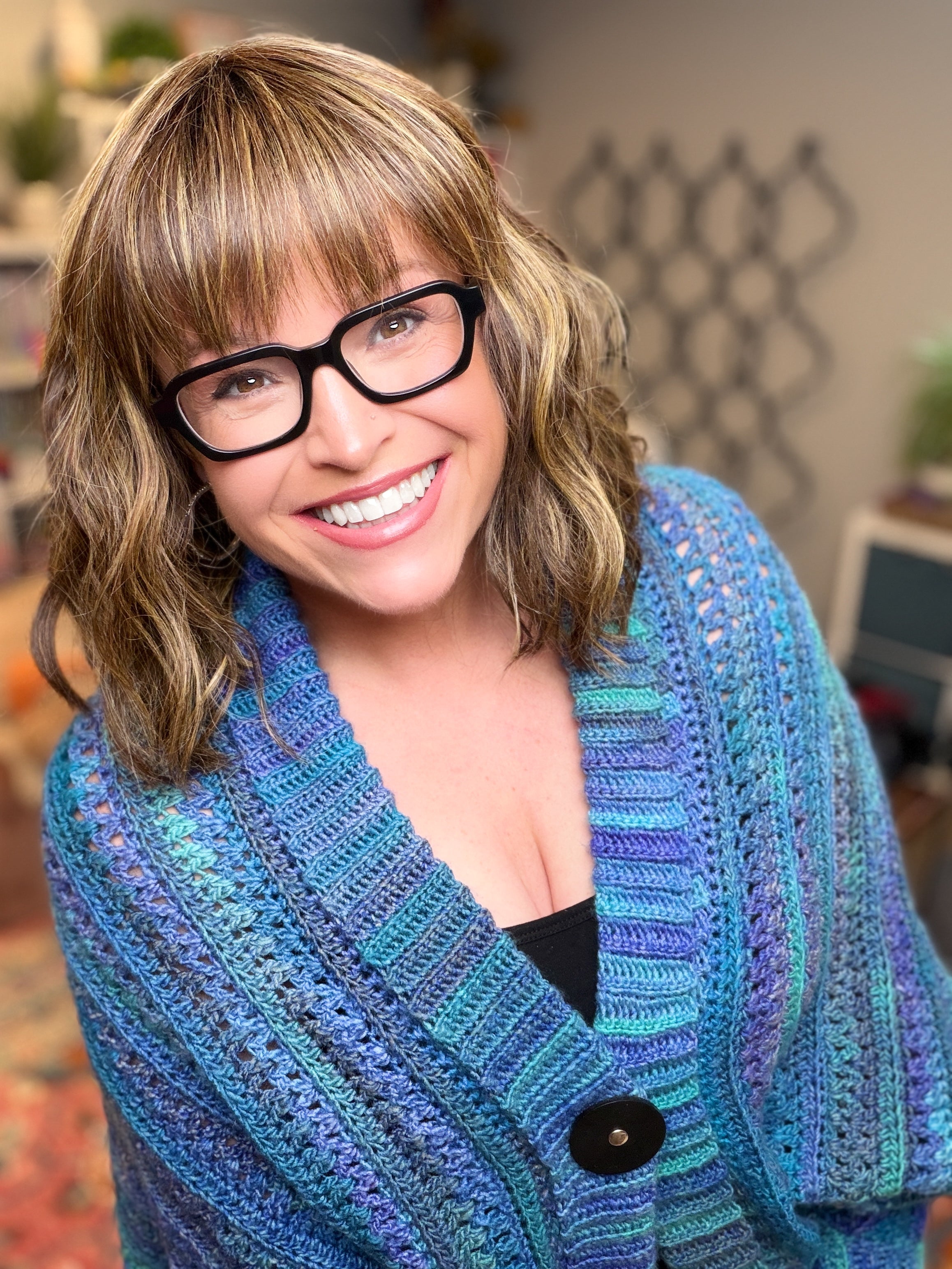 A smiling woman with wavy brown hair and glasses stands indoors in soft light, wearing the “You Are Valued Crochet Cocoon Cardigan,” which features a textured stitch pattern in blue and purple hues as she looks at the camera.