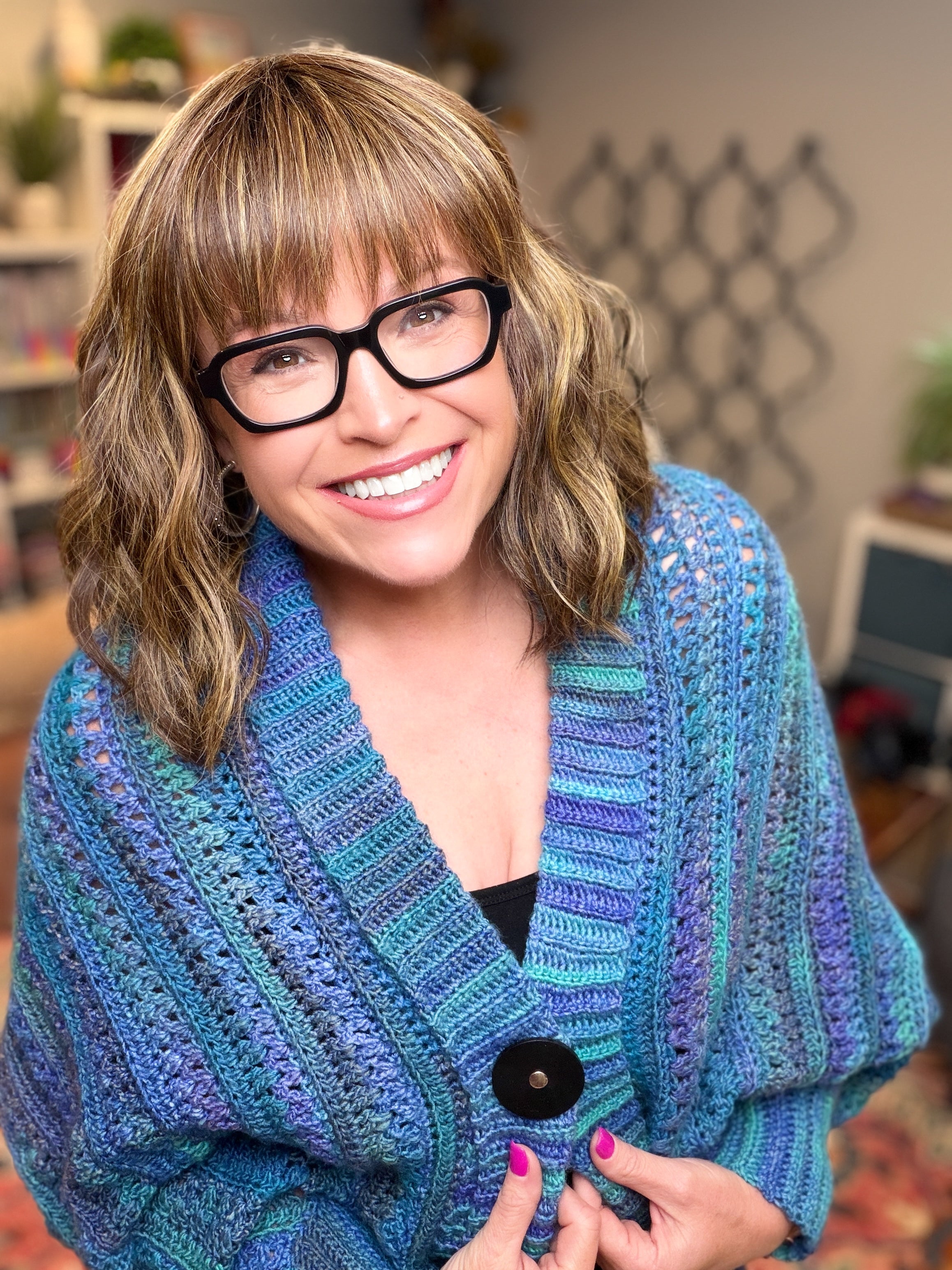 A smiling woman with glasses and shoulder-length hair wears a blue You Are Valued Crochet Cocoon Cardigan, showcasing its beautiful textured stitch pattern. Indoors, she holds the cozy oversized sweater closed, with bookshelves and home decor visible in the background.