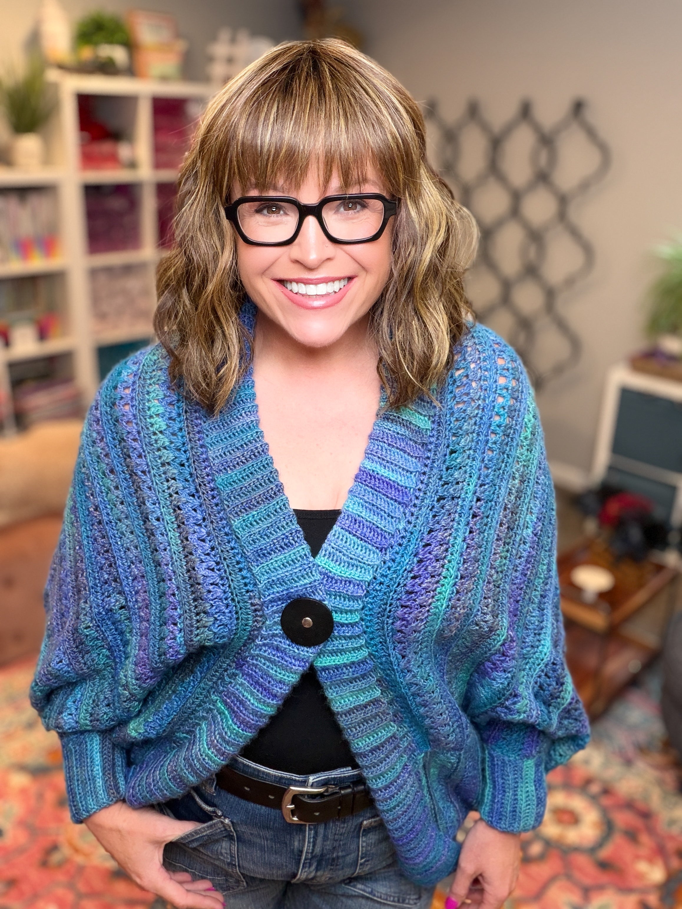 A smiling woman with shoulder-length hair and glasses wears the You Are Valued Crochet Cocoon Cardigan, an oversized blue and purple cardigan featuring a textured stitch pattern, while standing in a cozy, colorful room with shelves and plants in the background.