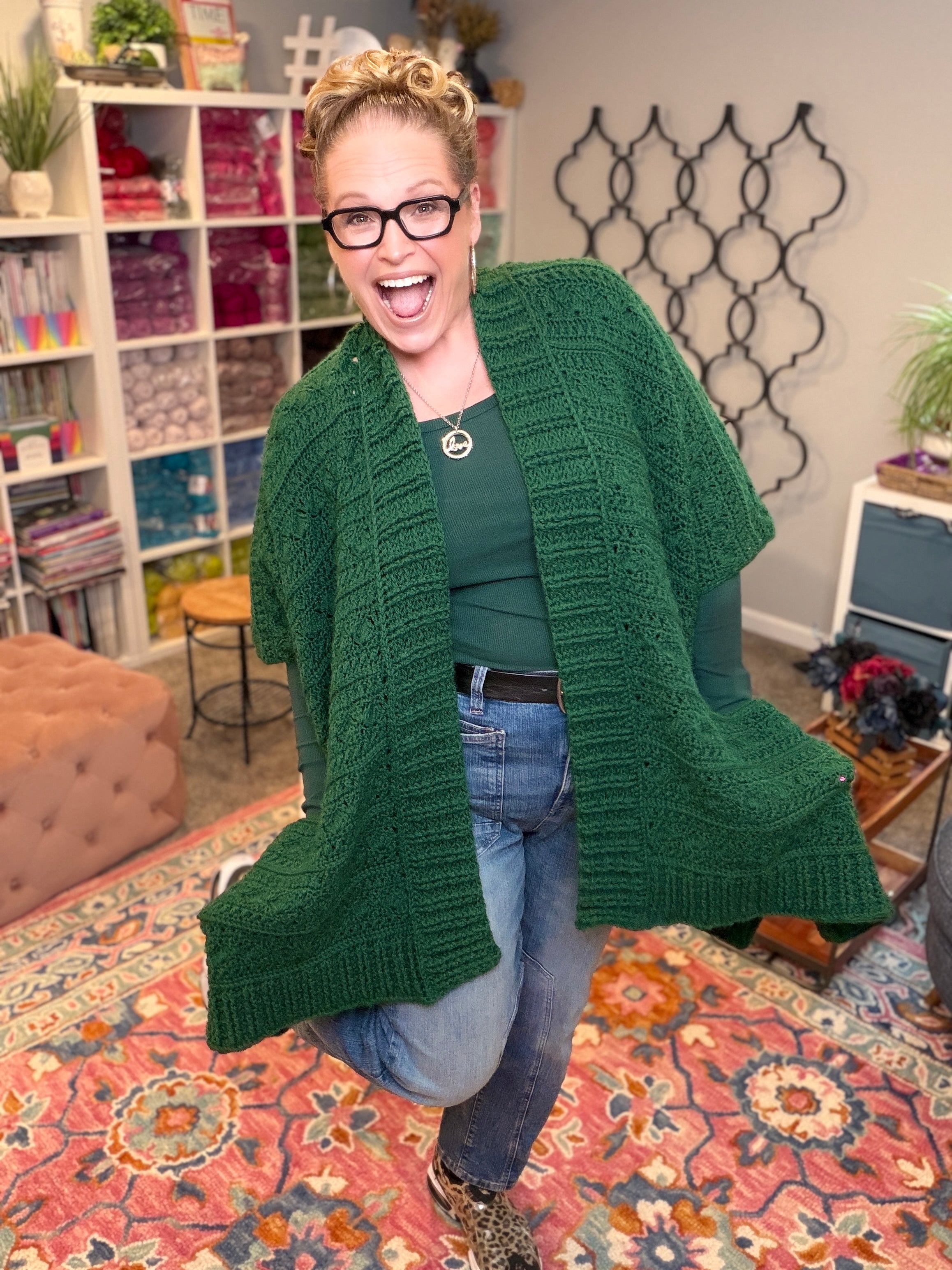 A smiling woman with blonde hair and glasses poses indoors, wearing a green Creekside Crochet Ruana crafted from Patons Classic Wool, paired with a green top, jeans, and leopard print shoes. She stands on a colorful rug with shelves and decor in the background.