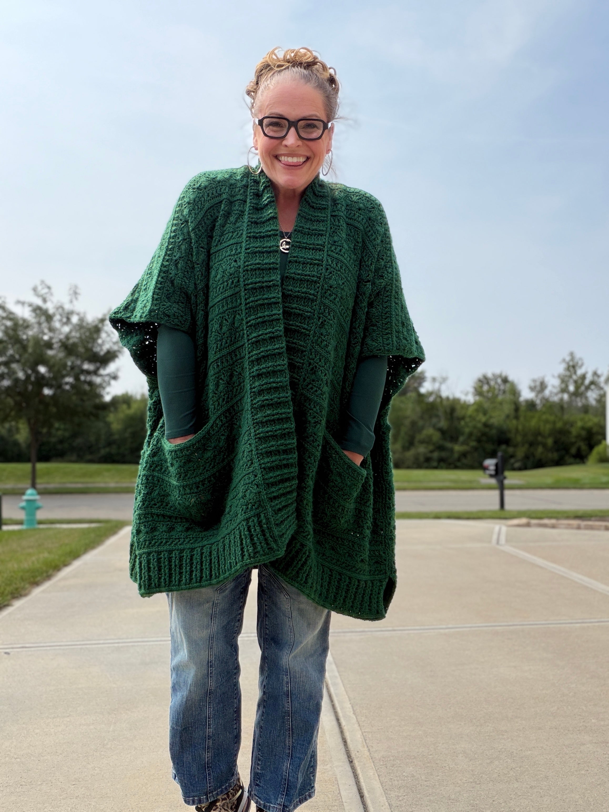 A smiling person wearing glasses, a green Creekside Crochet Ruana crafted from Patons Classic Wool, a green long-sleeve shirt, and blue jeans stands outdoors on a sidewalk with grass, trees, and a blue sky in the background.