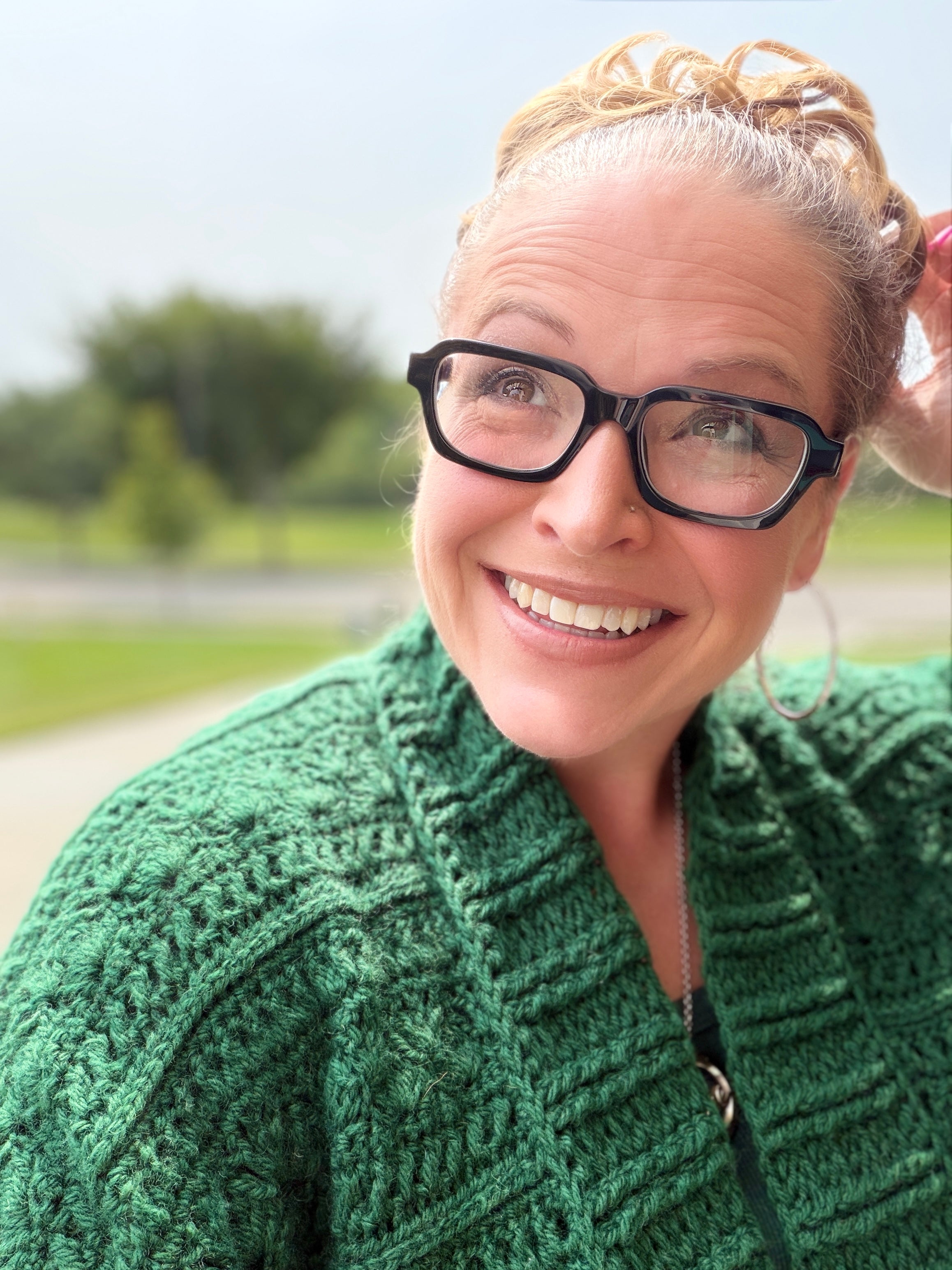 A smiling person with glasses and a green knitted sweater stands outdoors, draped in an oversized wrap made using the Creekside Crochet Ruana Pattern, with trees and a road blurred in the background.