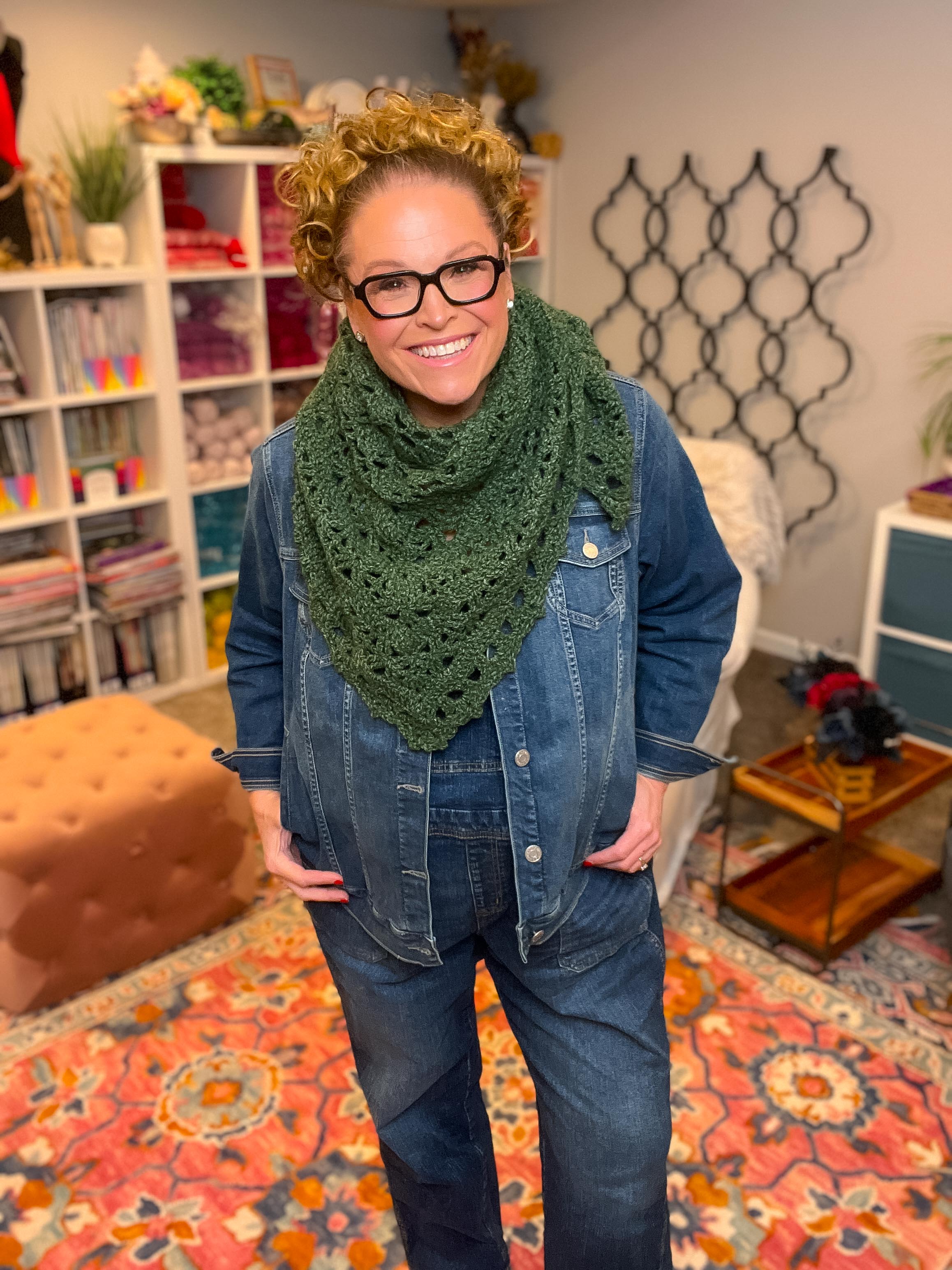 Woman wearing a green scarf and denim jacket in a room with a bookshelf and decorative items.